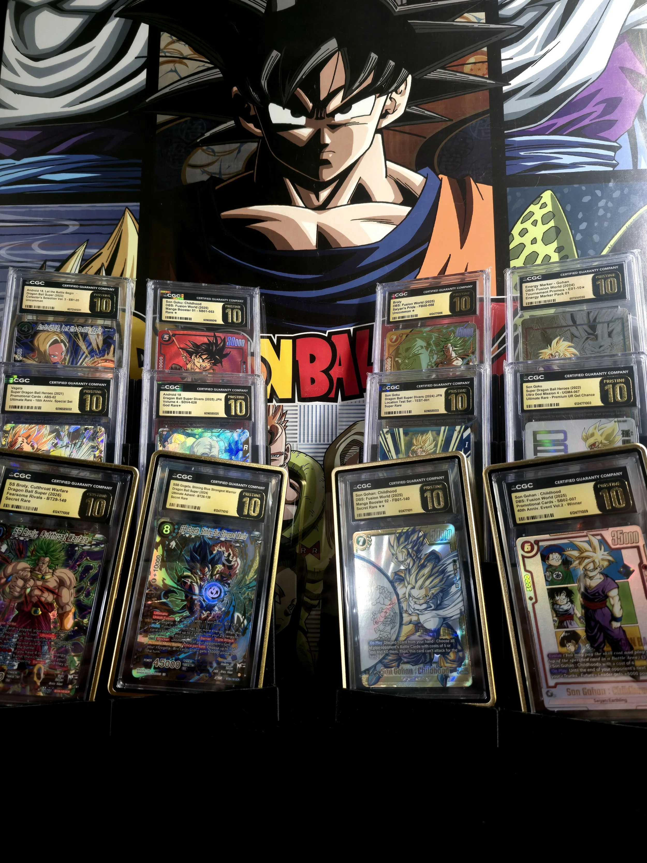 Display of various collectible Dragon Ball Super trading cards featuring characters like Son Goku, Vegeta, and Broly, arranged in front of a large Dragon Ball Z poster.