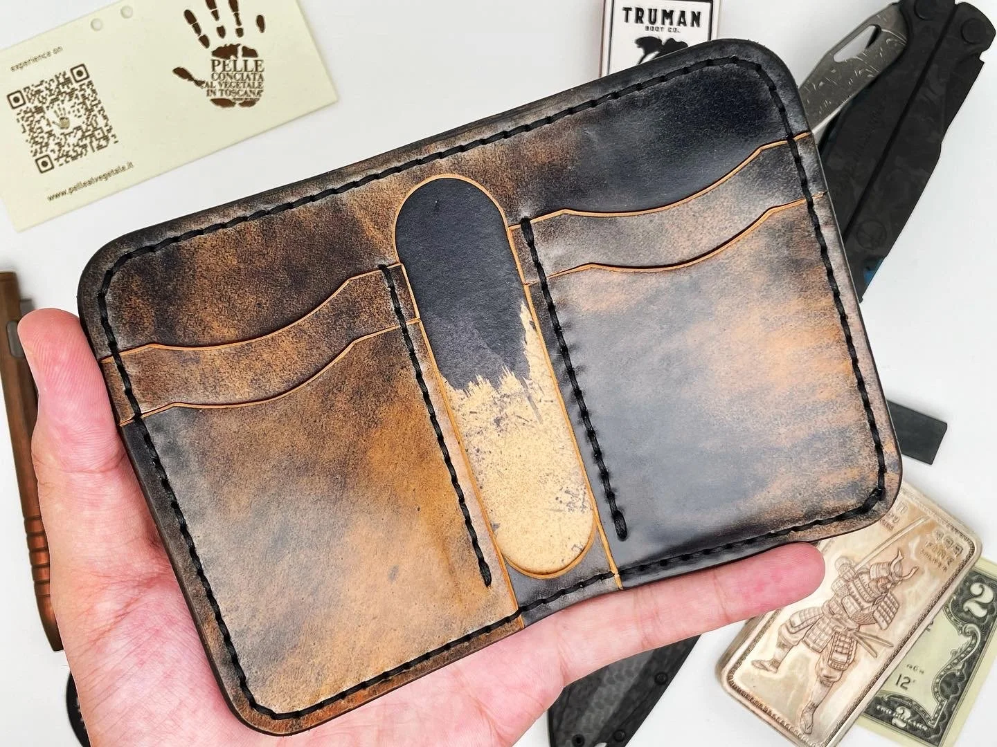 A hand holding a brown and black leather wallet with card slots. In the background, there is a hunting knife, a business card, a knife packaging, and some paper currency.
