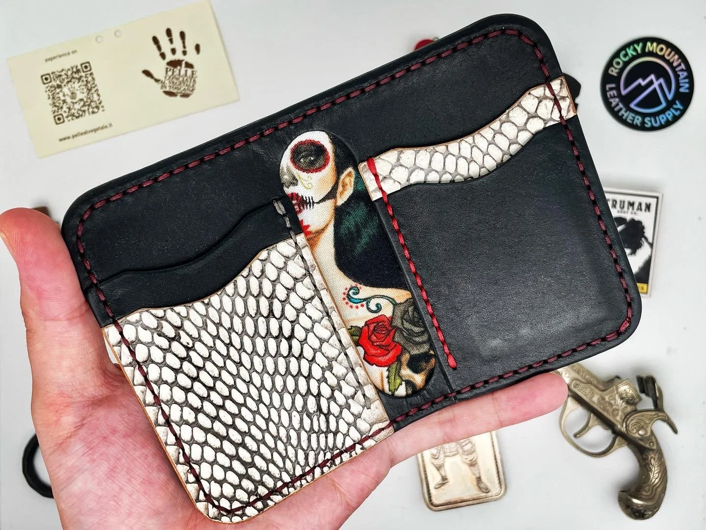 A black leather wallet with red stitching, decorated with skull and snake patterns, and a colorful tattoo-style skull and rose design.