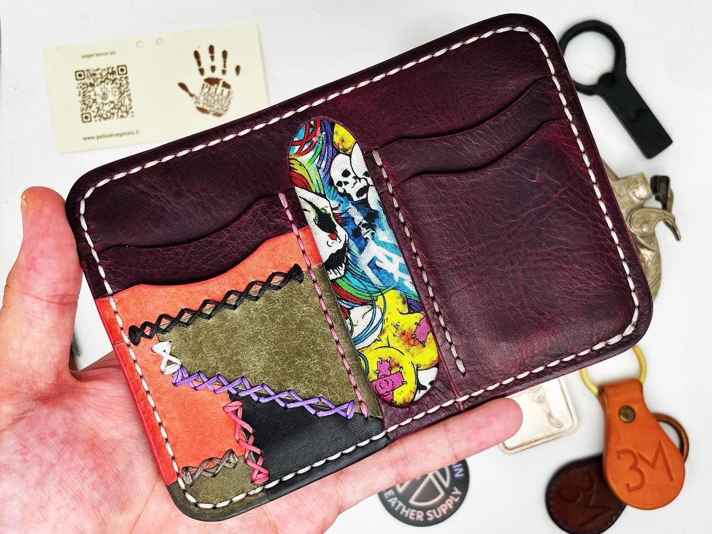 A colorfully stitched leather wallet with multiple card slots, held in a person's hand, with a business card and various keychains in the background.