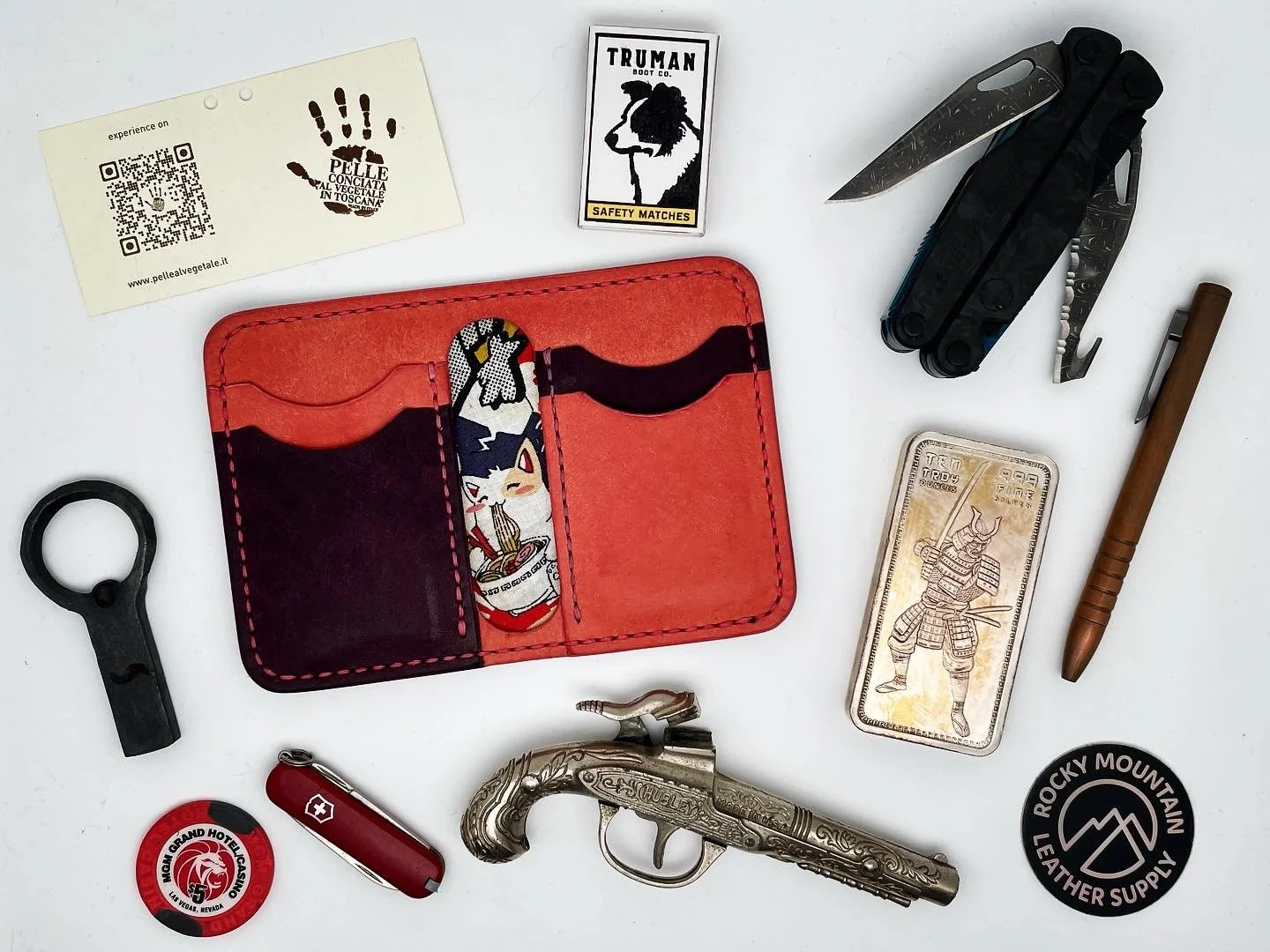 Collection of various items on a white surface, including a red wallet with a cat sticker, a multi-tool, a mini gun, a Swiss Army knife, a metallic lighter, a black and white patch, a vintage-style card with a warrior, a pen, a key tool, a small tin,