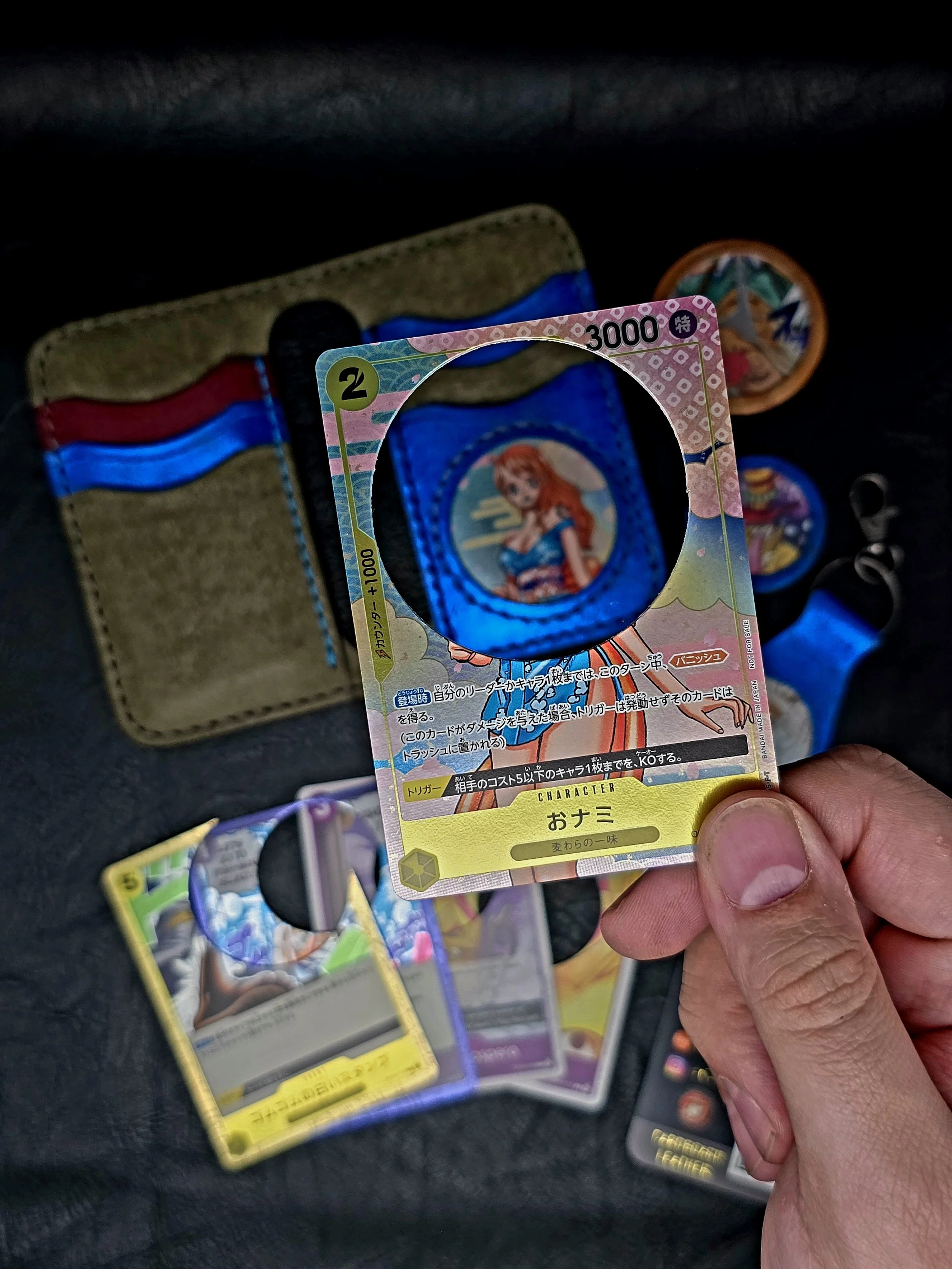 Close-up of a hand holding a trading card from a Japanese card game, depicting a female character with red hair. Other cards and a cloth wallet are visible in the background.