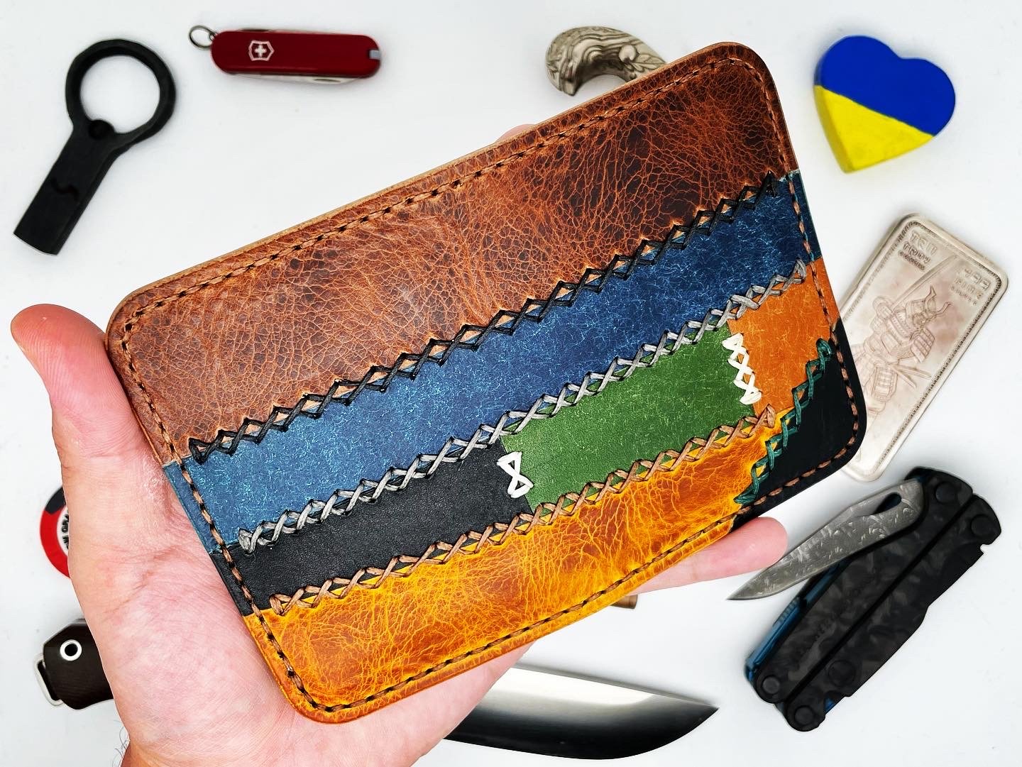 Colorful leather wallet with zigzag stitching, surrounded by a Swiss Army knife, a heart-shaped item painted with Ukraine's flag colors, a spiral knife, and knives on a white surface.