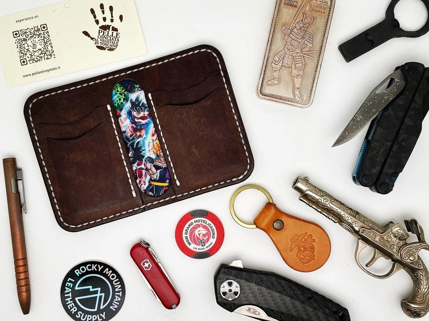 Collection of miscellaneous items on a white surface, including a leather wallet with colorful fabric interior, a pen, a poker chip, a Swiss Army knife, a leather keychain, a vintage pistol, a pocket knife, a multi-tool, a pair of scissors, a copper 