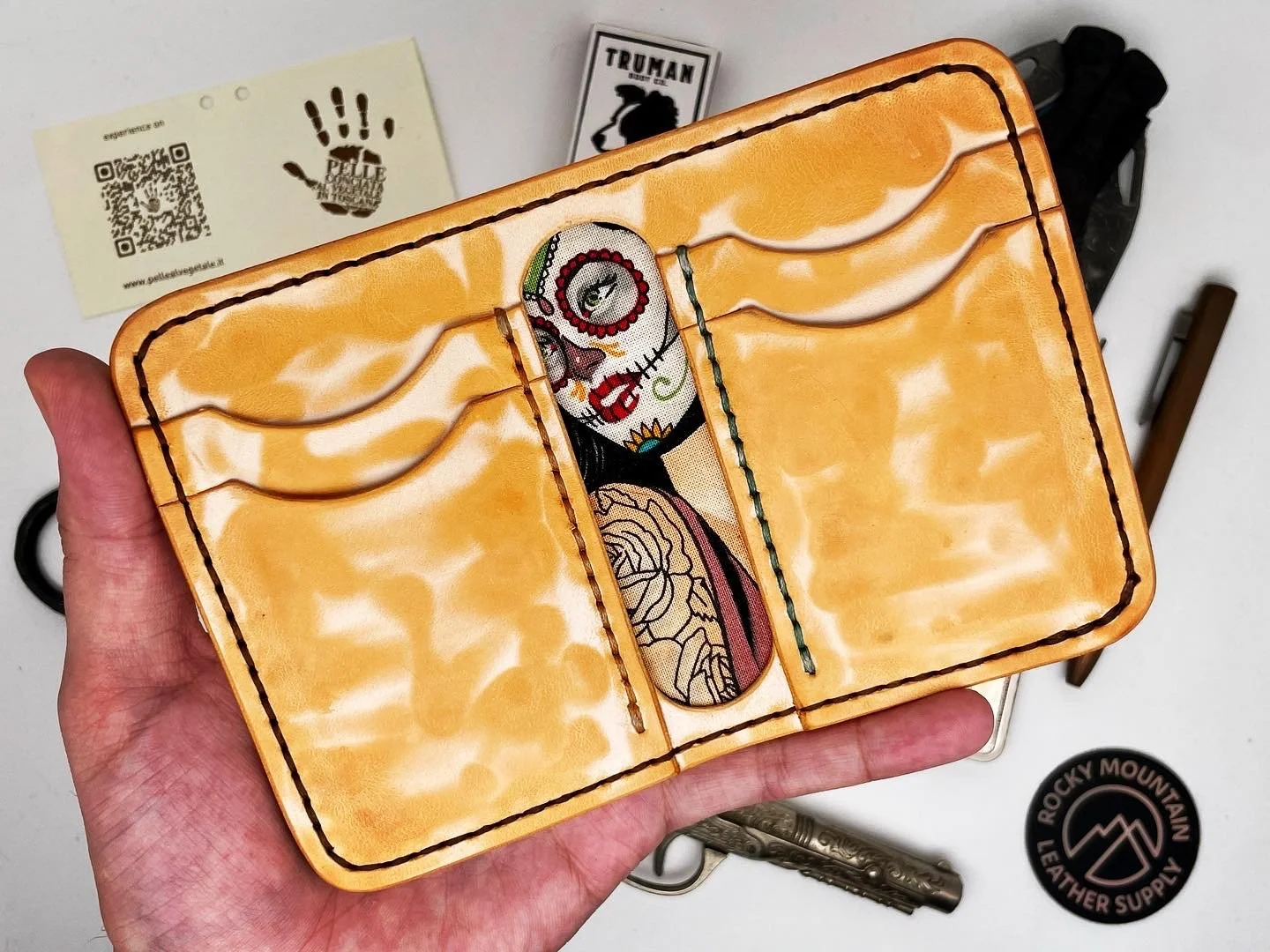 Hand holding a leather wallet or clutch with a colorful embroidered design of a woman, featuring a tattoo on her chest and face makeup, placed on a white surface with various accessories around it.