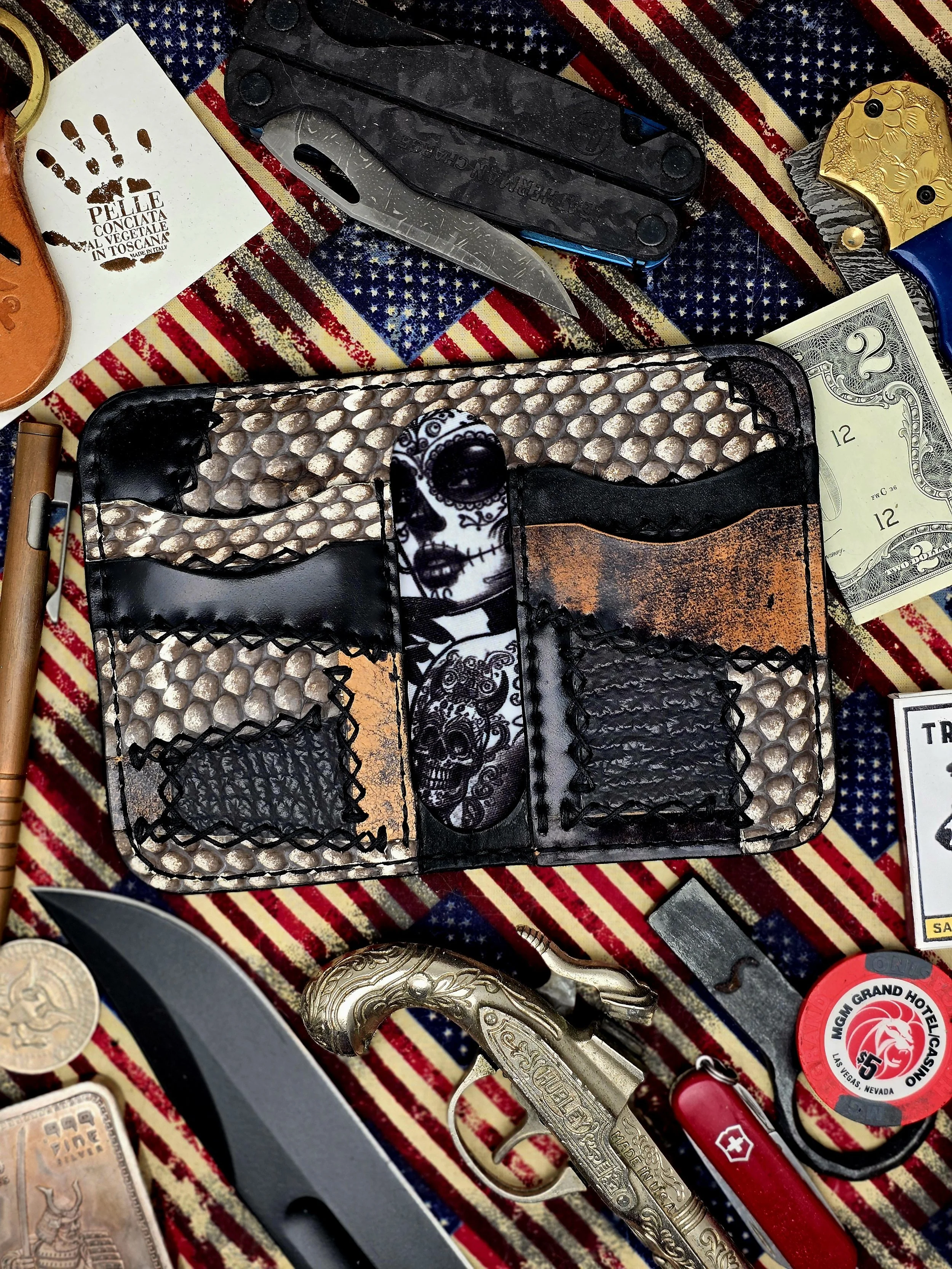 Collection of personal items scattered on an American flag-themed fabric, including a colorful briefcase with skull and skeleton design, a folding knife, a smaller pocket knife, a red Swiss Army knife, a black and silver knife, a gold-colored pistol-