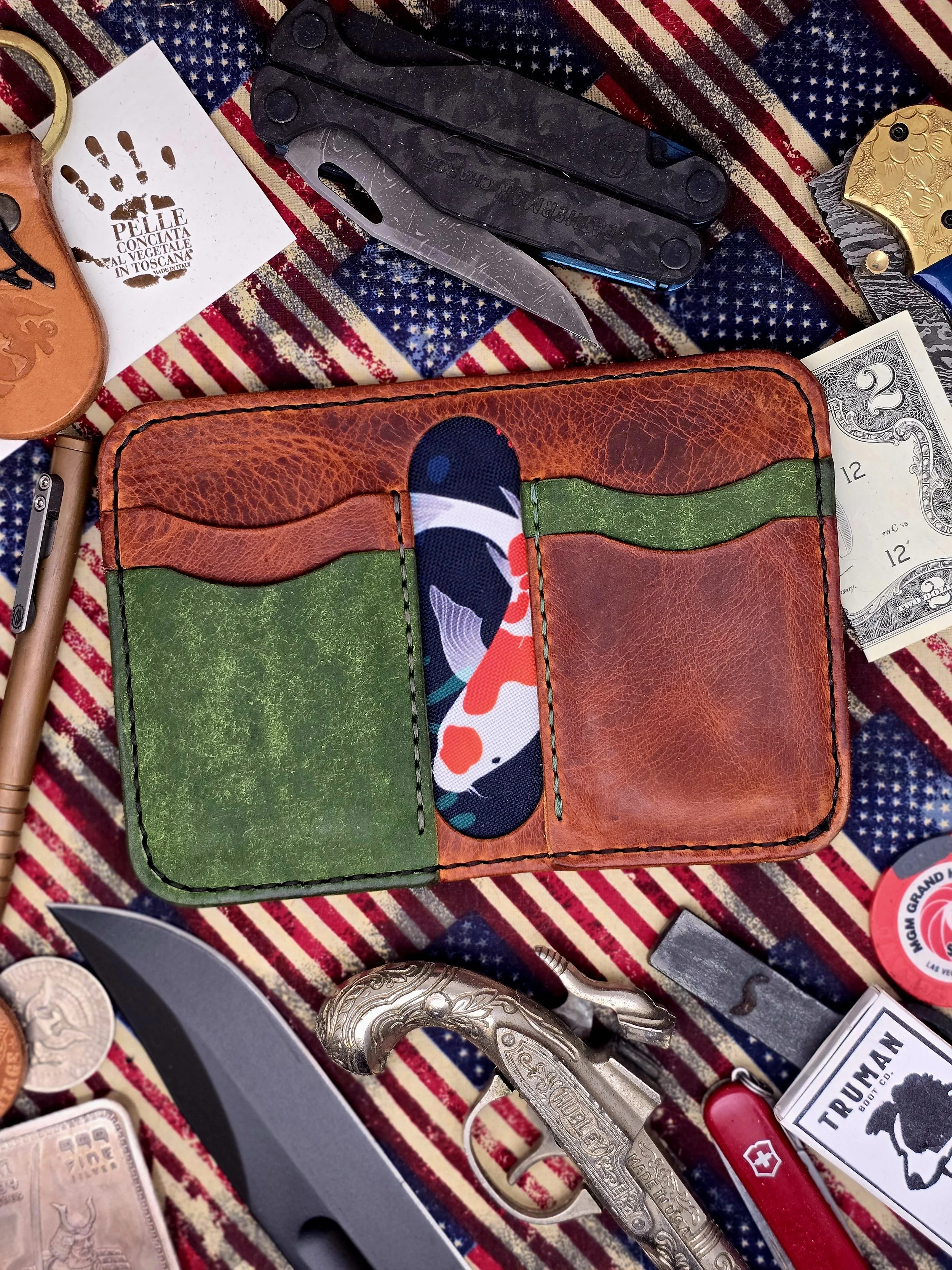 A leather wallet with red, green, and camouflage patterns placed on a patriotic fabric background, surrounded by various knives, a pistol, a gun-shaped bottle opener, a Swiss Army knife, coins, and stickers.