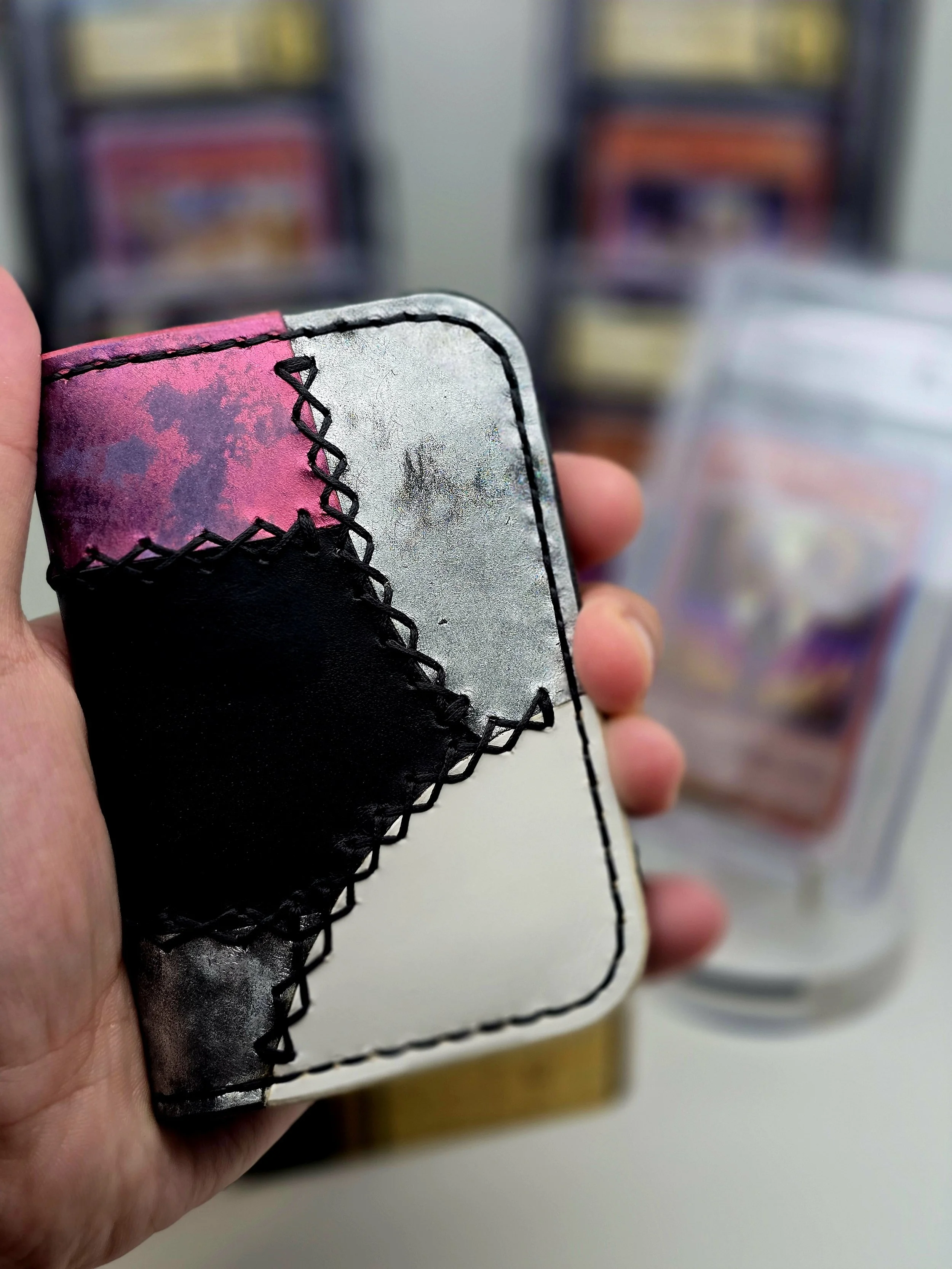 A hand holding a small patchwork leather wallet with pink, black, white, and metallic silver pieces, stitched with black thread, with a blurred background of vending machines.