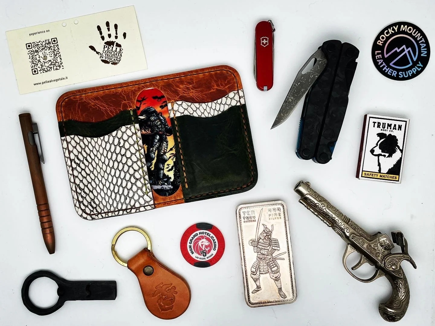 A collection of miscellaneous items on a white surface, including a patterned leather wallet, a red Swiss Army knife, a multi-tool, a small safety matchbox, a vintage-style pistol, a keychain with a leather tag, a poker chip, a card with a samurai il