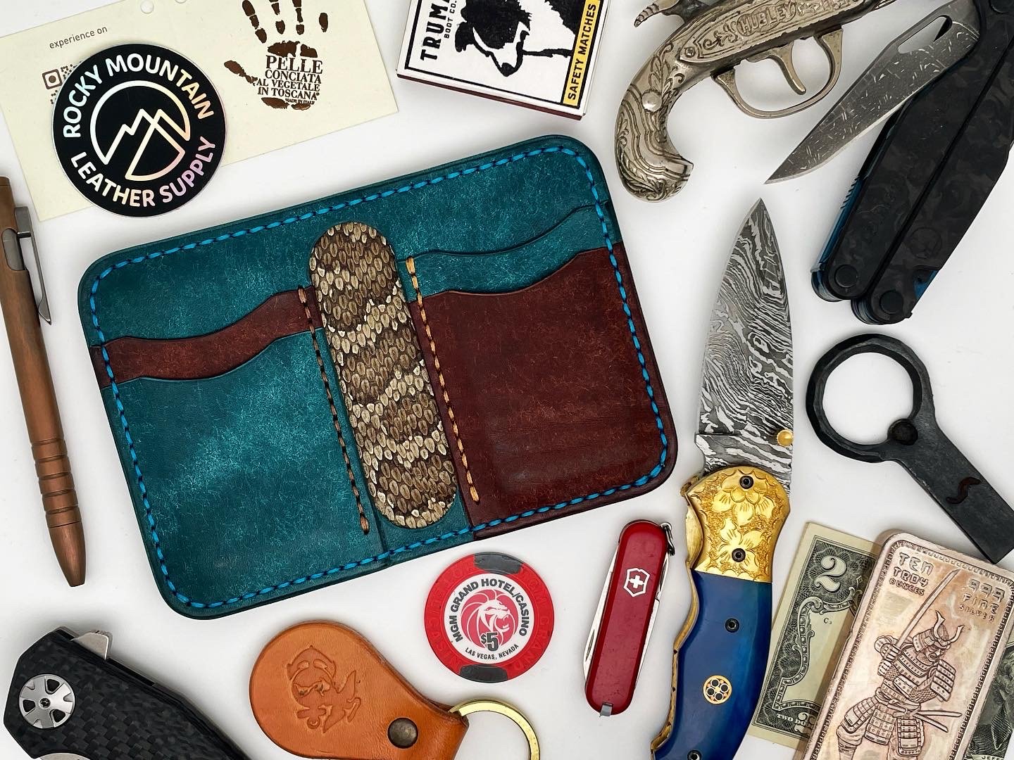 Collection of everyday items including a leather wallet, a pocket knife with a decorative gold design, a Swiss army knife, folded cash, a USB drive, a leather keychain, a poker chip, a multi-tool, a small folding knife with a Damascus-style blade, an