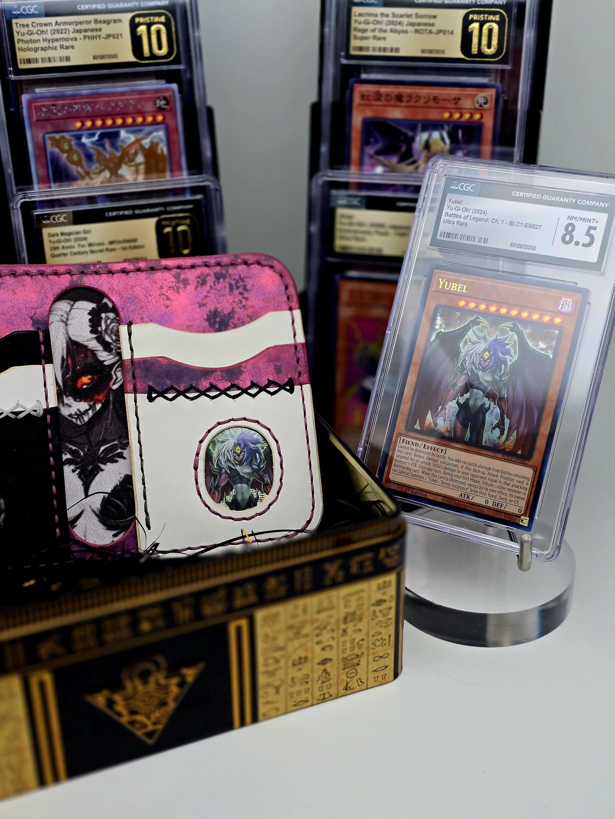 Photo of multiple collectible trading cards, including a Yu-Gi-Oh! Yubel card graded 8.5 in a plastic slab, a pink and black card holder with a Yubel card inside, and other cards in the background with grading labels.