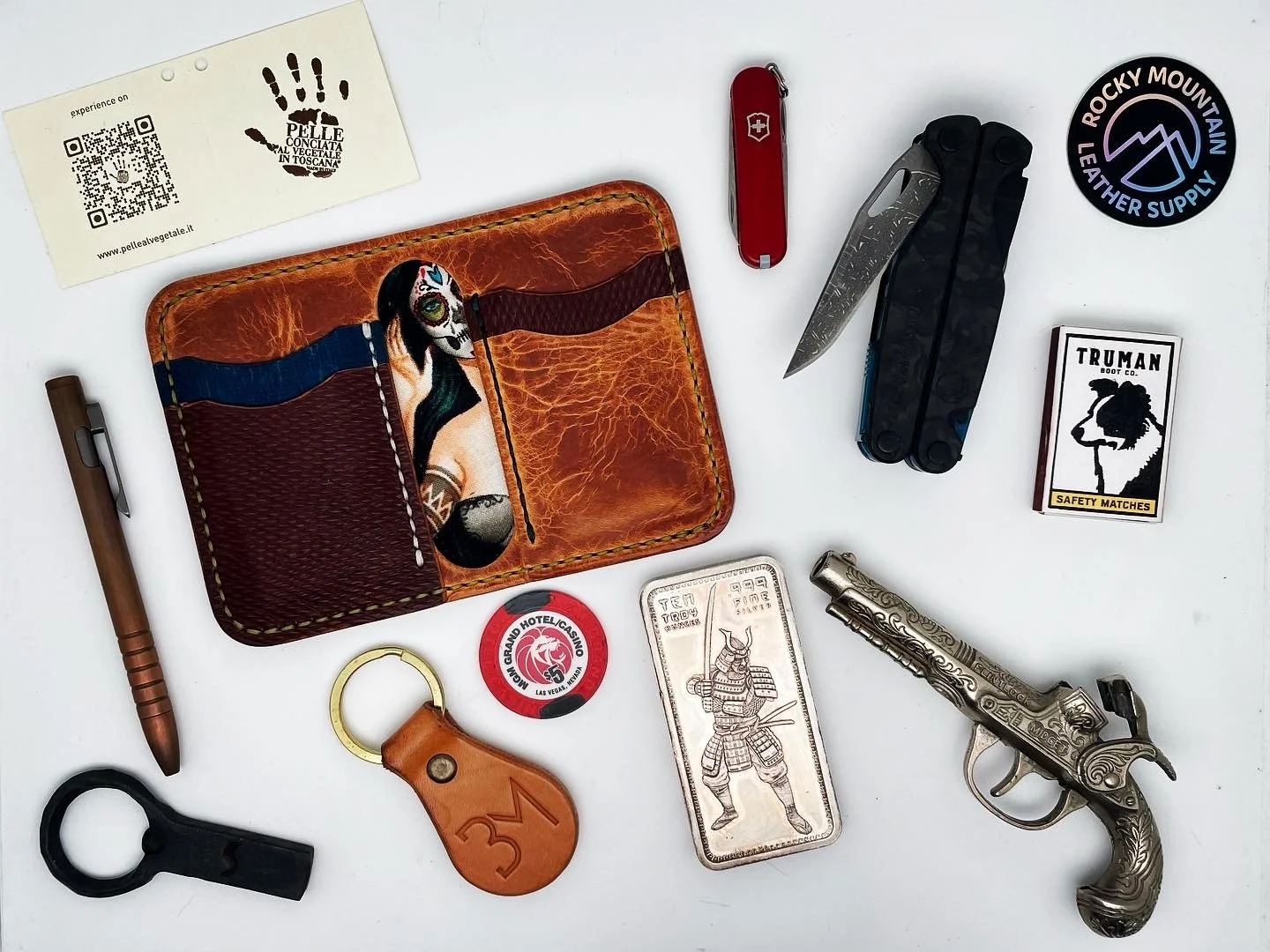 Collection of various items including a leather wallet with a Day of the Dead skeleton design, a Swiss Army knife, a smaller pocket knife, a vintage-style pistol, a matchbox, a keyring with a leather tag, a poker chip, a matchbook, a leather-cased li