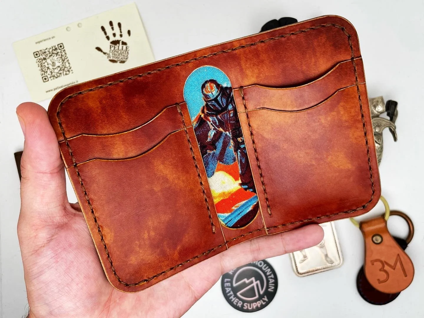 A brown leather wallet with multiple card slots, featuring a colorful sticker of an astronaut on the front. There's a black and white patch with a mountain logo, a small silver case, a brown leather keychain, and a business card with a QR code and a 