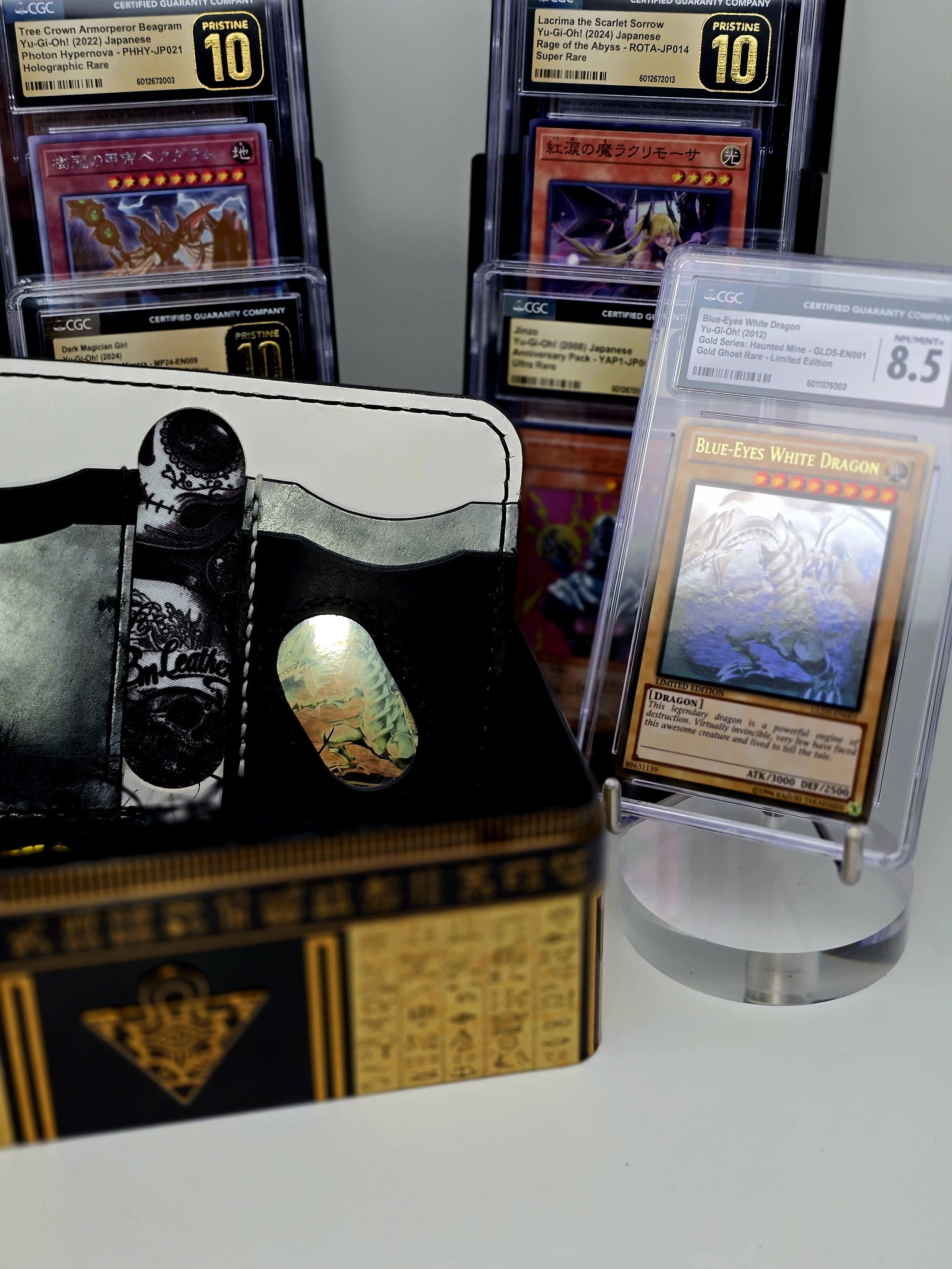 A collection of Yu-Gi-Oh! trading cards, including an item titled 'Blue-Eyes White Dragon' graded 8.5, displayed on a stand, with some cards in protective cases in the background.