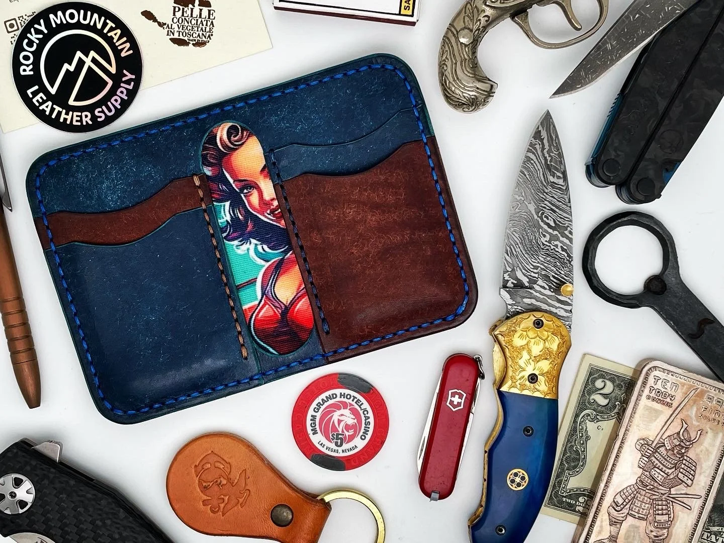 Collection of various items including a leather wallet with a pin-up girl pattern, emergency knife, pocket knife with blue and gold handle, flashlight, Swiss Army knife, dollar bills, poker chip, sunglasses case, and USB flash drive.