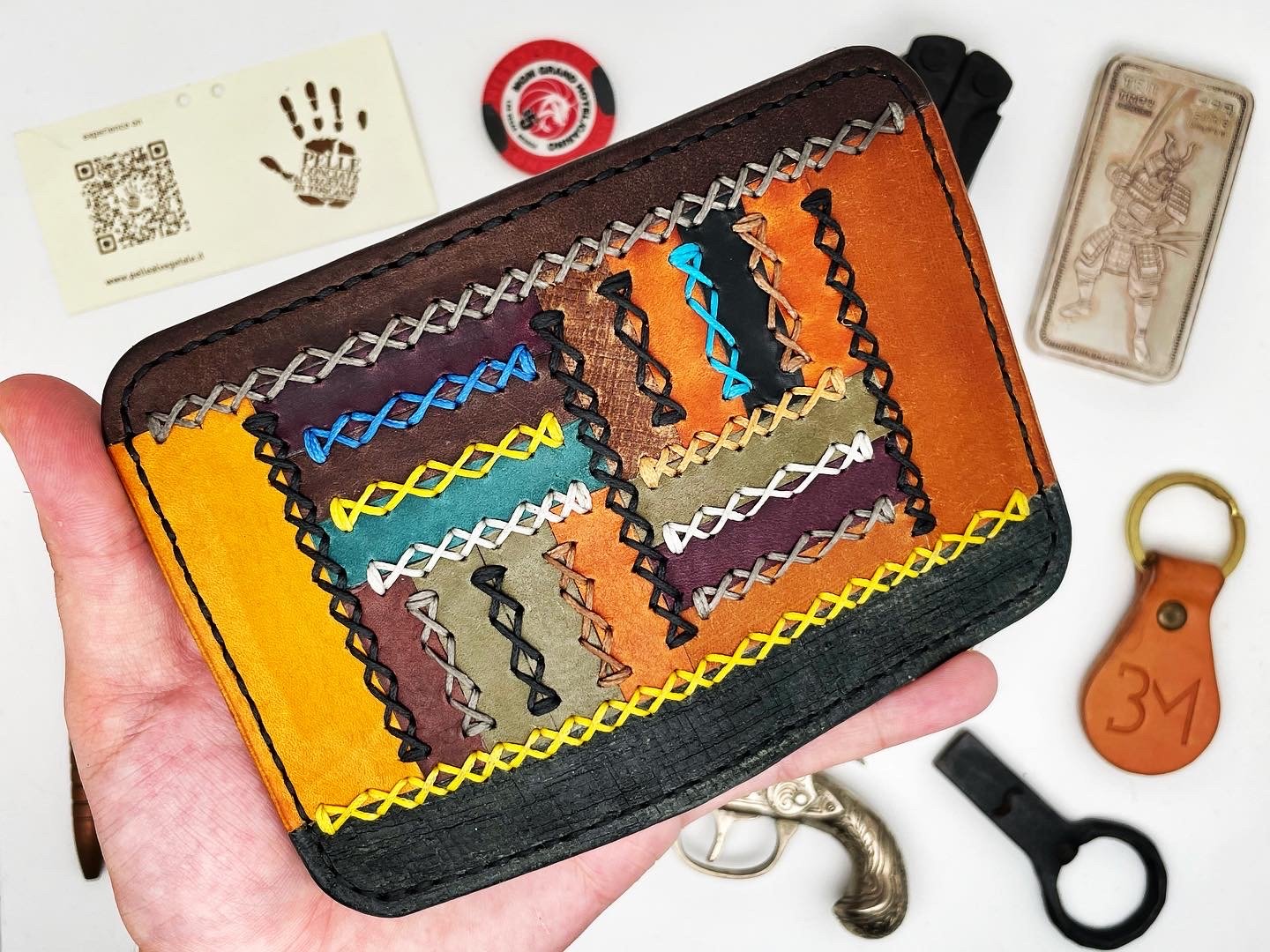 Colorful handcrafted leather wallet with decorative stitching and patchwork design, held in a person's hand. Various metal and leather accessories are visible on a white surface around it.