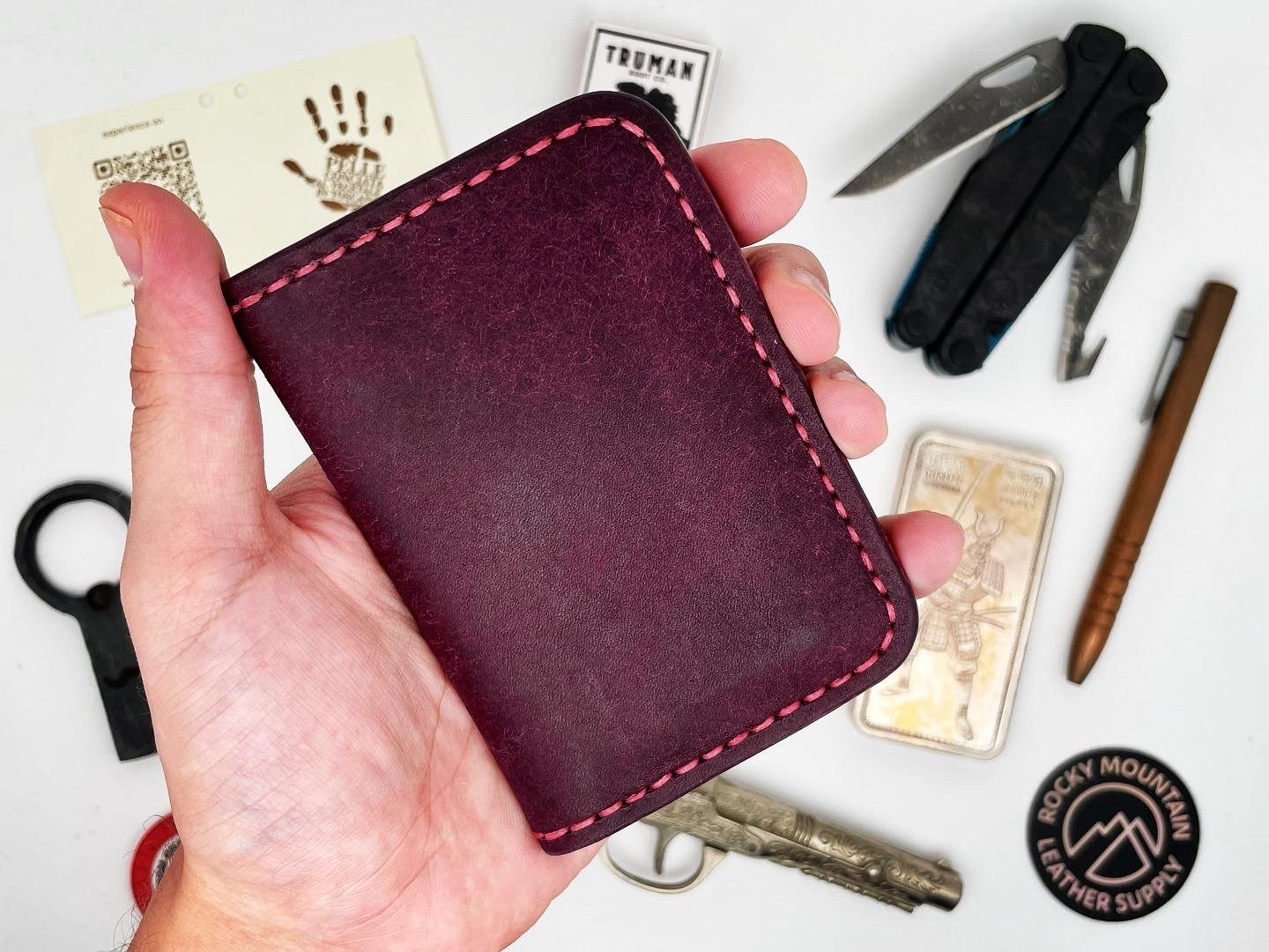 A hand holding a maroon wallet with pink stitching, surrounded by various tools and items including a multi-tool, a small knife, a card with a handprint, and other miscellaneous objects.