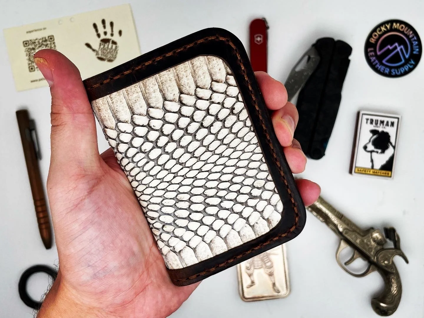 Hand holding a wallet with a snake skin pattern. Various items on a white surface include a knife, a revolver, a Swiss Army knife, patches, stickers, and a card.