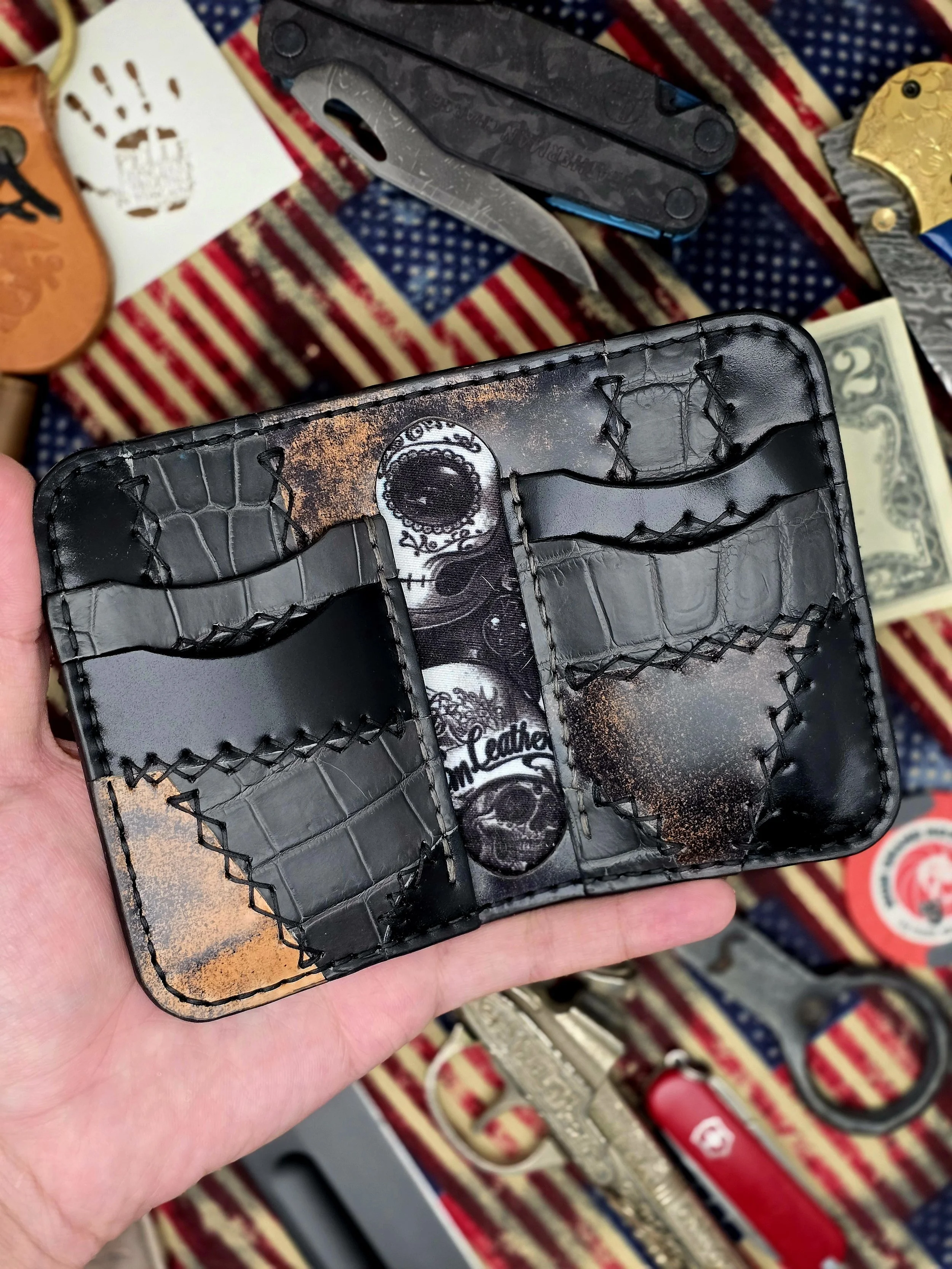 Black leather wallet with skull design inside, held in hand, displaying card slots and interior details, with various tools and items on a striped fabric background.