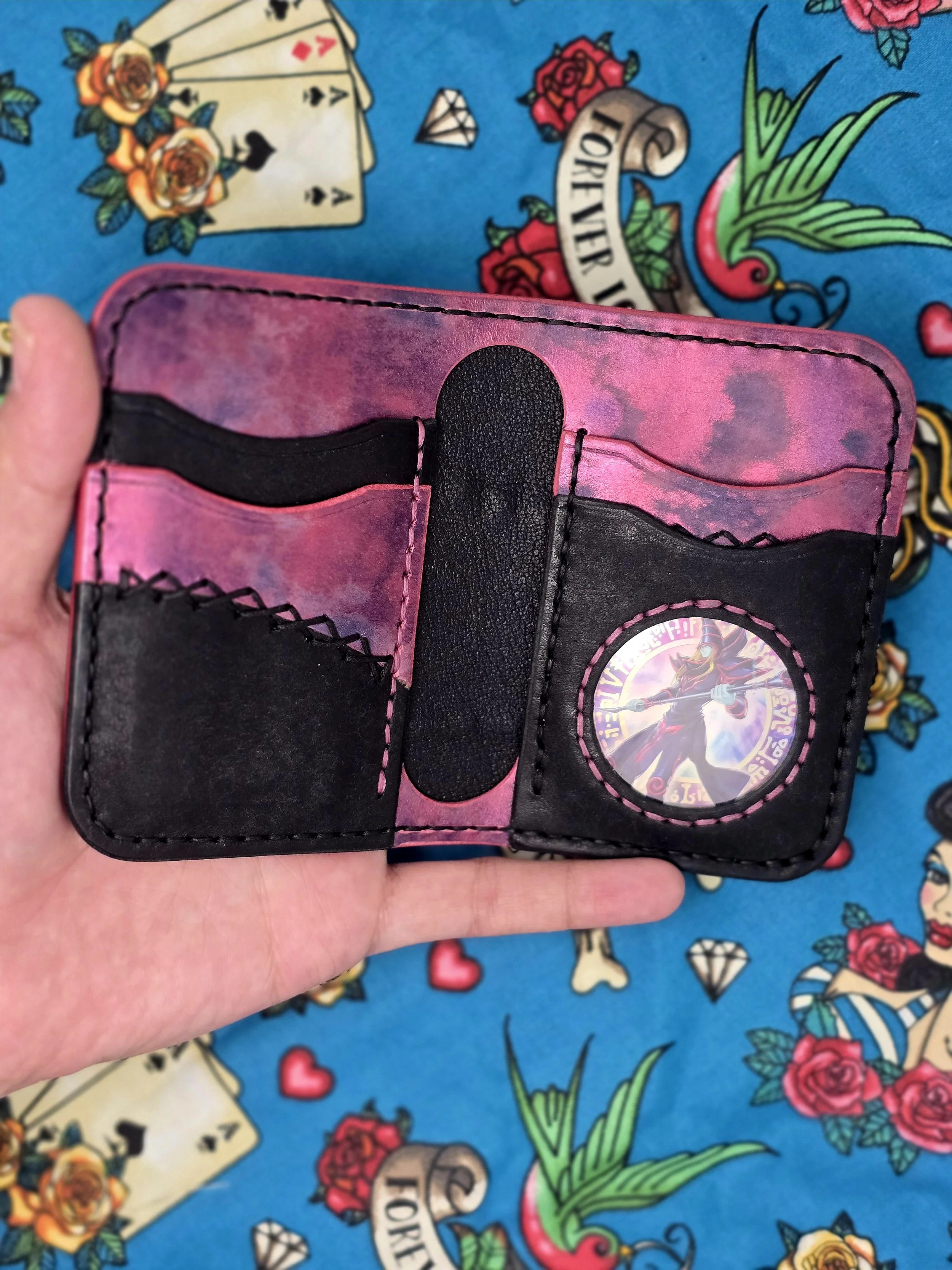 Open black and pink wallet with card slots and a holographic sticker featuring a character with a sword, held against a colorful tattoo-themed fabric background.
