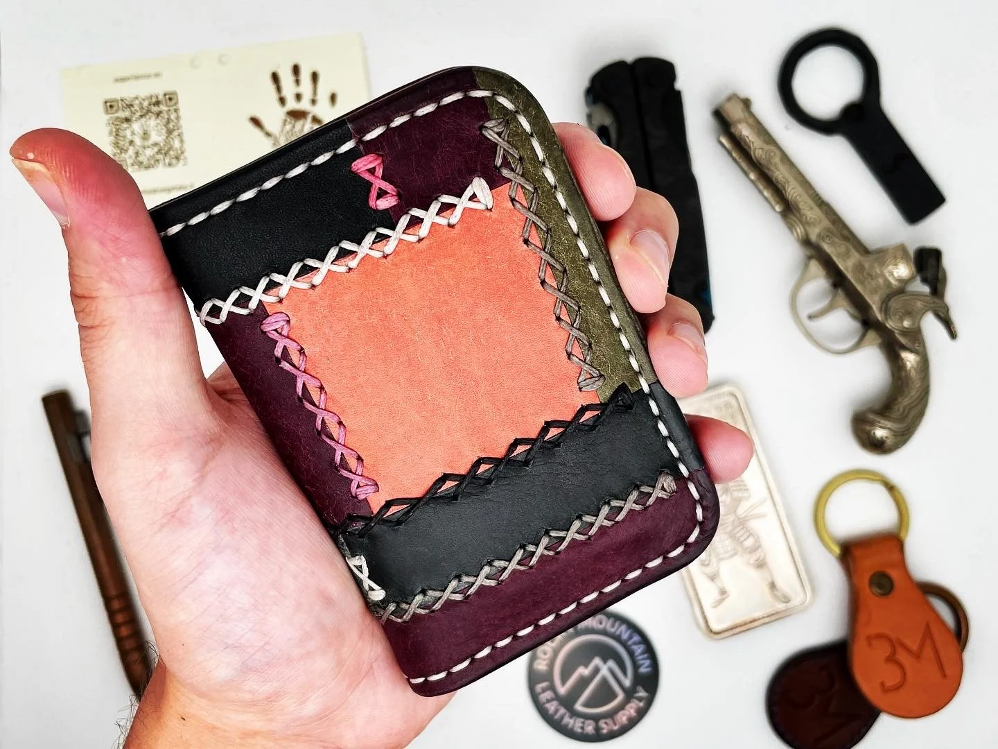 A hand holds a colorful, patchwork leather wallet with decorative stitching. Surrounding objects include a vintage gun, two keychains, a badge, a sticker, a QR code card, and a lighter.