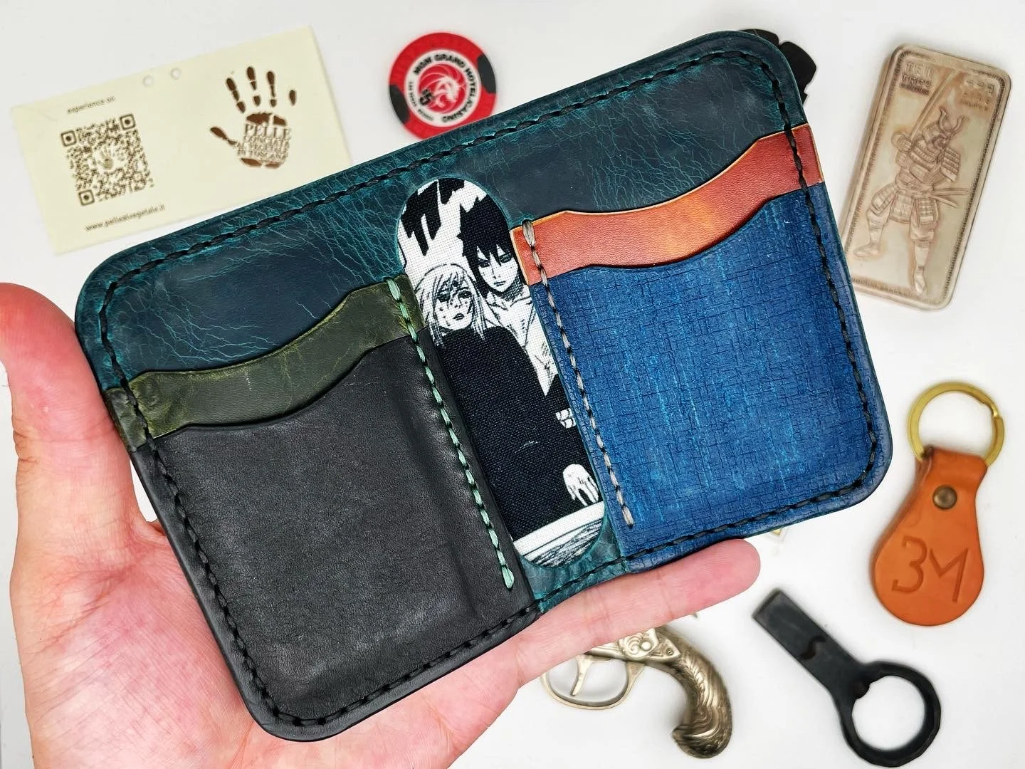 Open colorful leather wallet containing a comic strip illustration, surrounded by various items including a business card, a round sticker, a banknote, a leather keychain, a toy gun, a key, and a metal figurine.