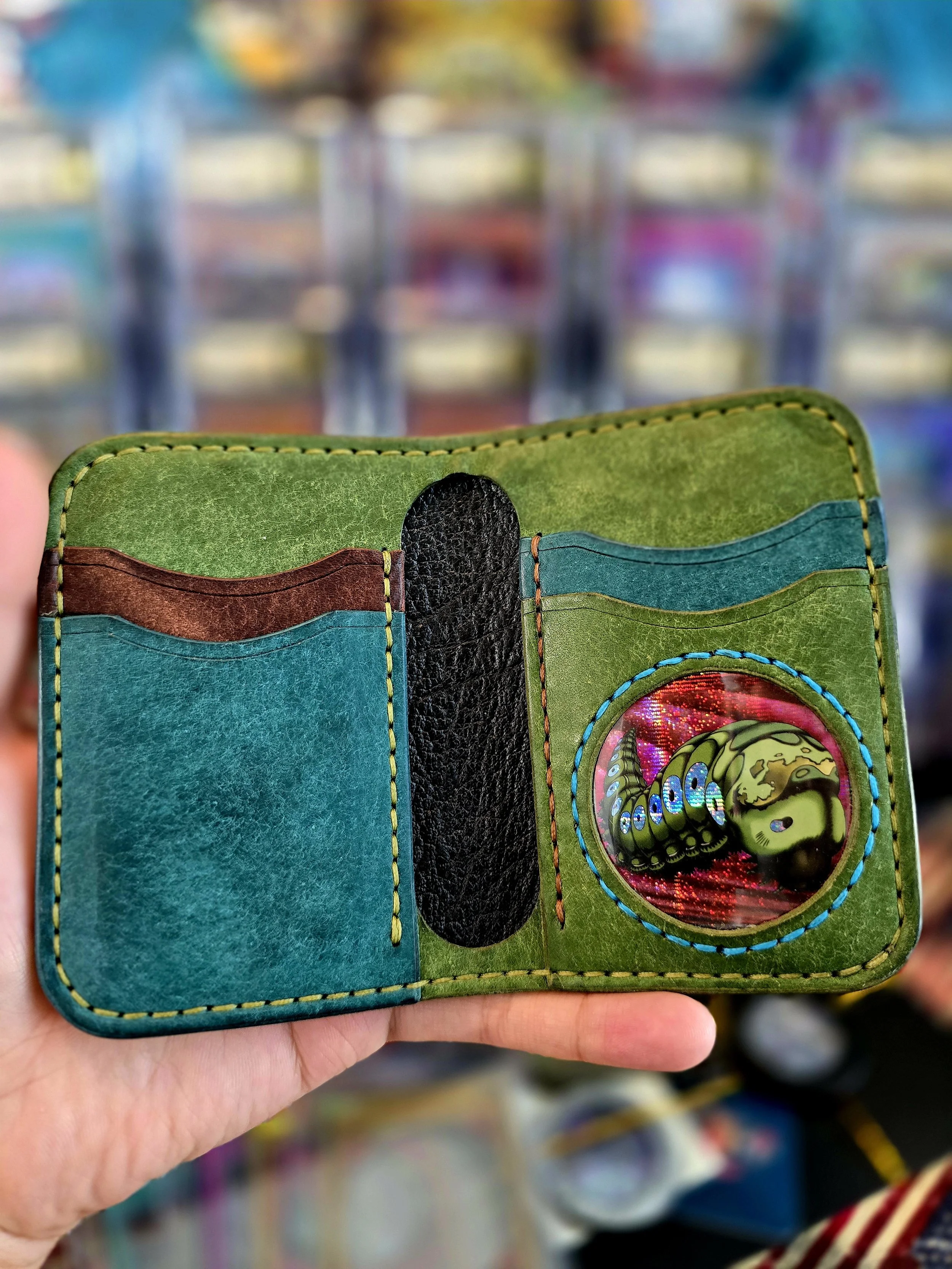 A hand holding a colorful, decorated wallet with multiple card slots and a circular sticker or button design inside. The wallet has green, blue, brown, and black sections with visible stitching.