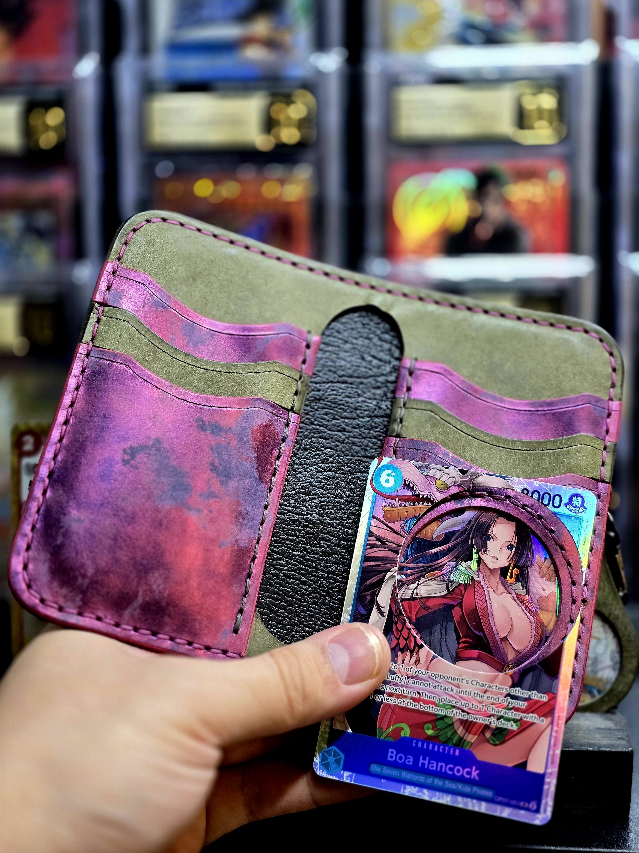 Open wallet with pink, purple, and green accents, holding trading cards, including one of Boa Hancock from One Piece, in front of a background with vending machines or arcade games.
