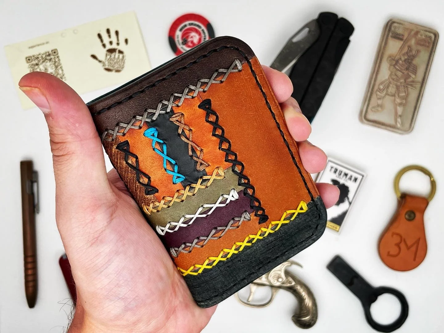 Hand holding a colorful, stitched leather wallet with various items on a white surface in the background, including a keyring, a small gun-shaped object, a playing card, and other miscellaneous objects.