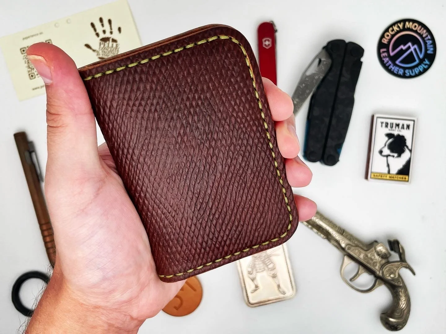 Hand holding a brown textured wallet with yellow stitching, surrounded by various items on a white surface including a Swiss Army knife, a black multitool, a Rocky Mountain leather supply badge, a matchbox with a dog head, a patch with a hand, a smal