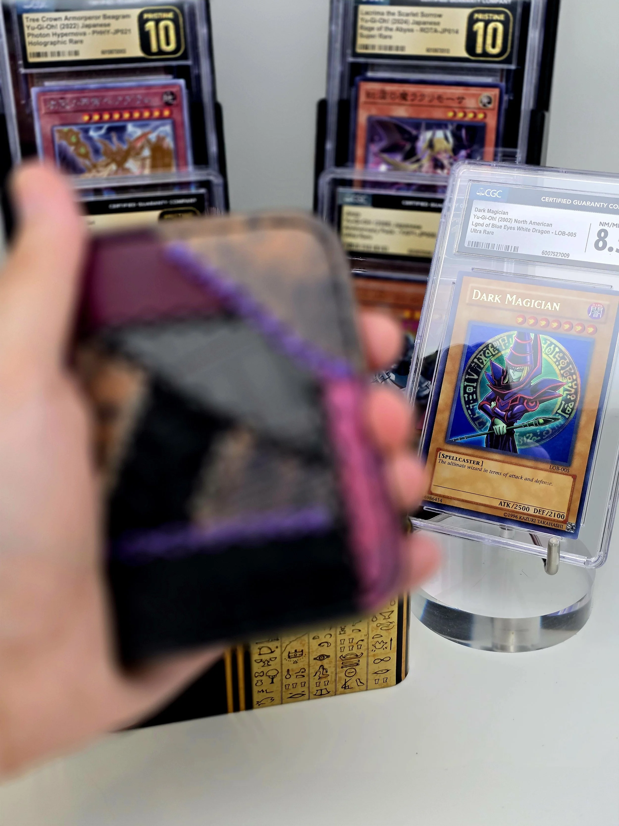 Close-up of a person's hand holding a small purple and black box, with a blurred collection of Yu-Gi-Oh! trading cards displayed in graded and ungraded cases in the background, including a Dark Magician card.