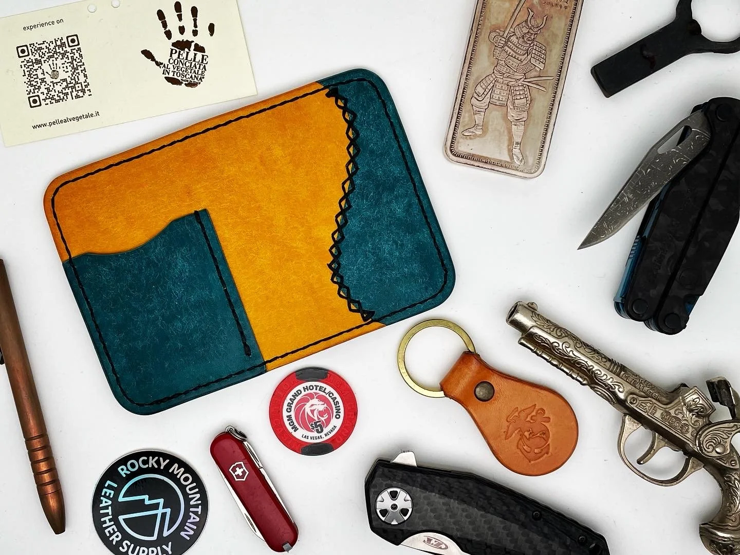 A collection of items including a colorful leather card holder, a business card from Pelle al Vegetale, a Japanese yen banknote depicting a samurai, a folding pocket knife, a silver decorative pistol, a round red token for the Grand Hotel Casino in L