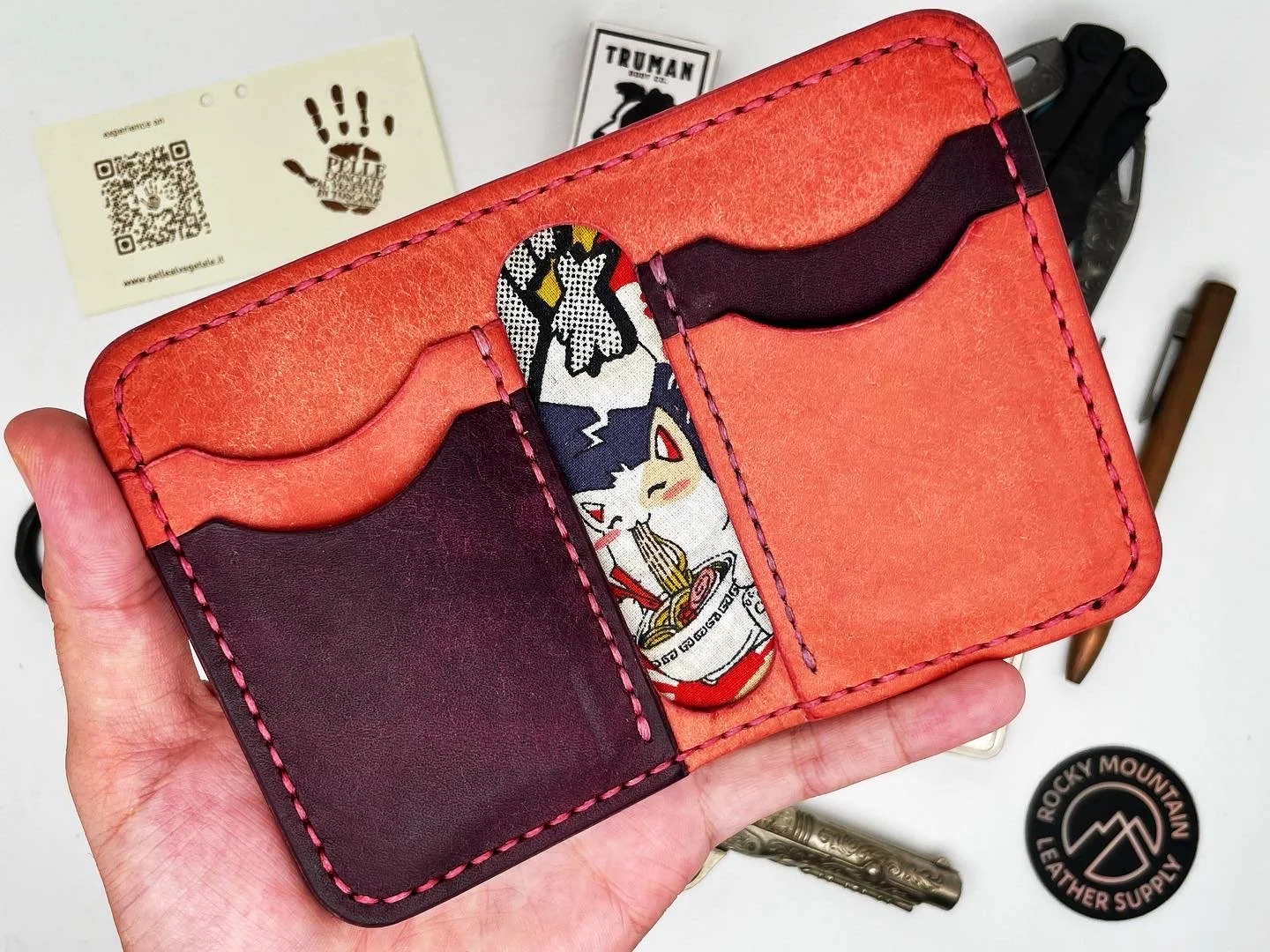 Open leather cardholder with multiple card slots, one featuring a colorful character fabric design, held in a person's hand. Items on a white surface include business cards, a small badge, and various tools.