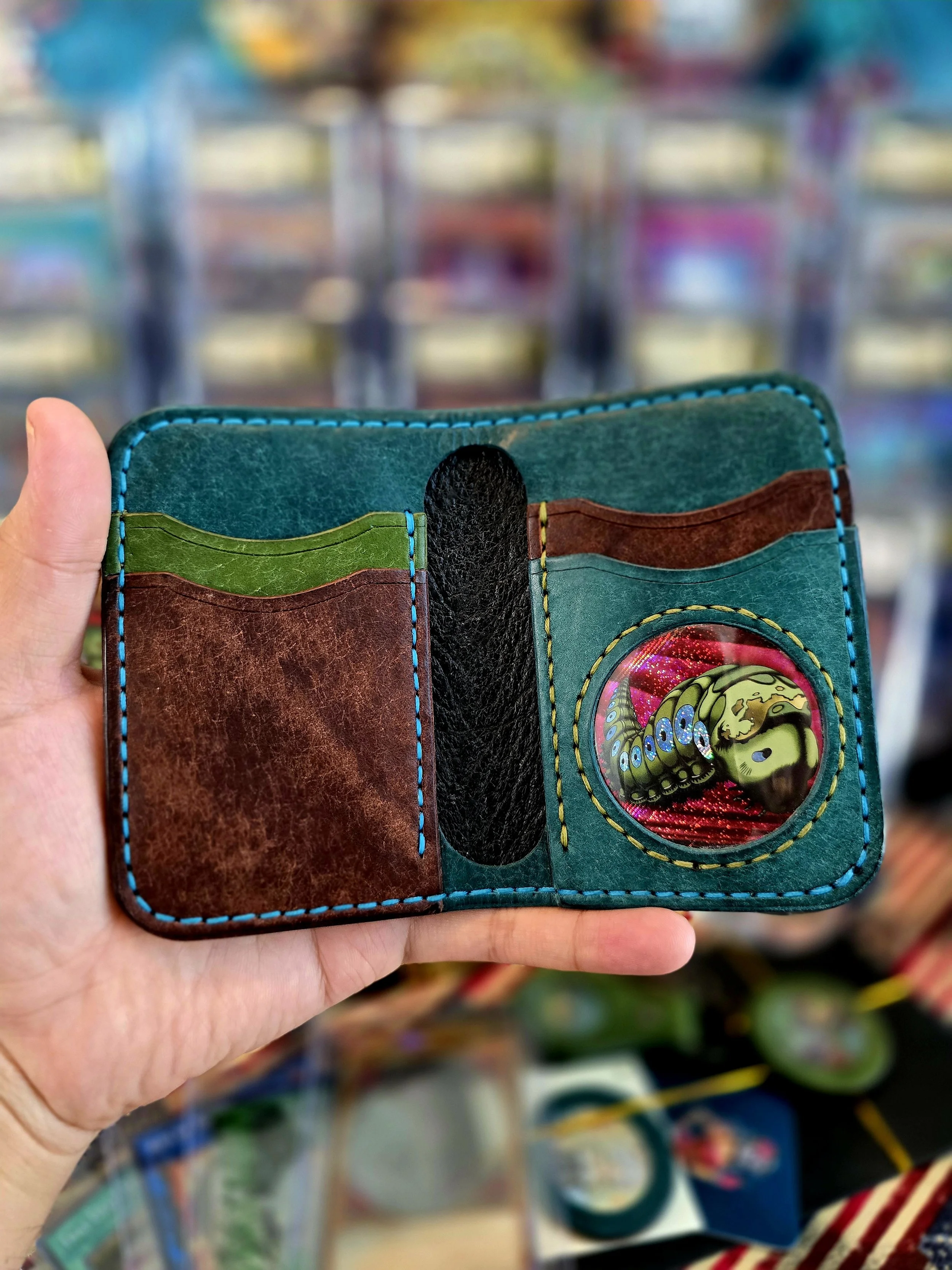 Colorful handmade leather wallet with card slots and a circular window showing a peacock feather and a yin-yang symbol, held in a person's hand against a blurred retail store background.