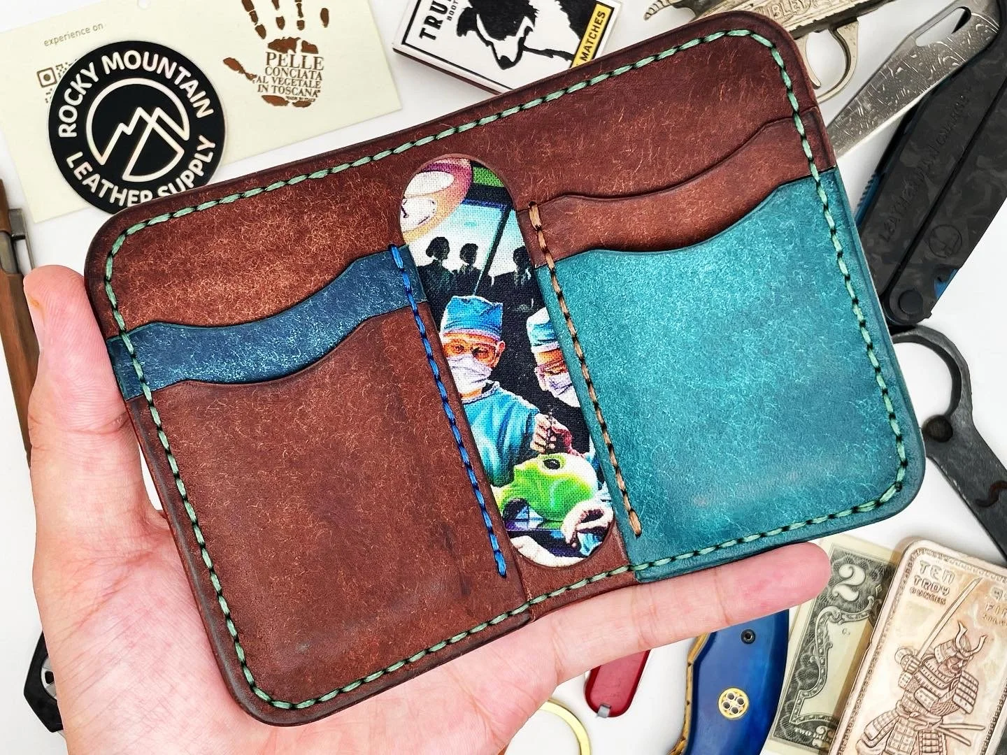 Hand holding a brown and blue leather wallet with colorful stitching and a money slot. Background includes a plastic toy gun, dollar bills, and stickers with outdoor and mountain themes.