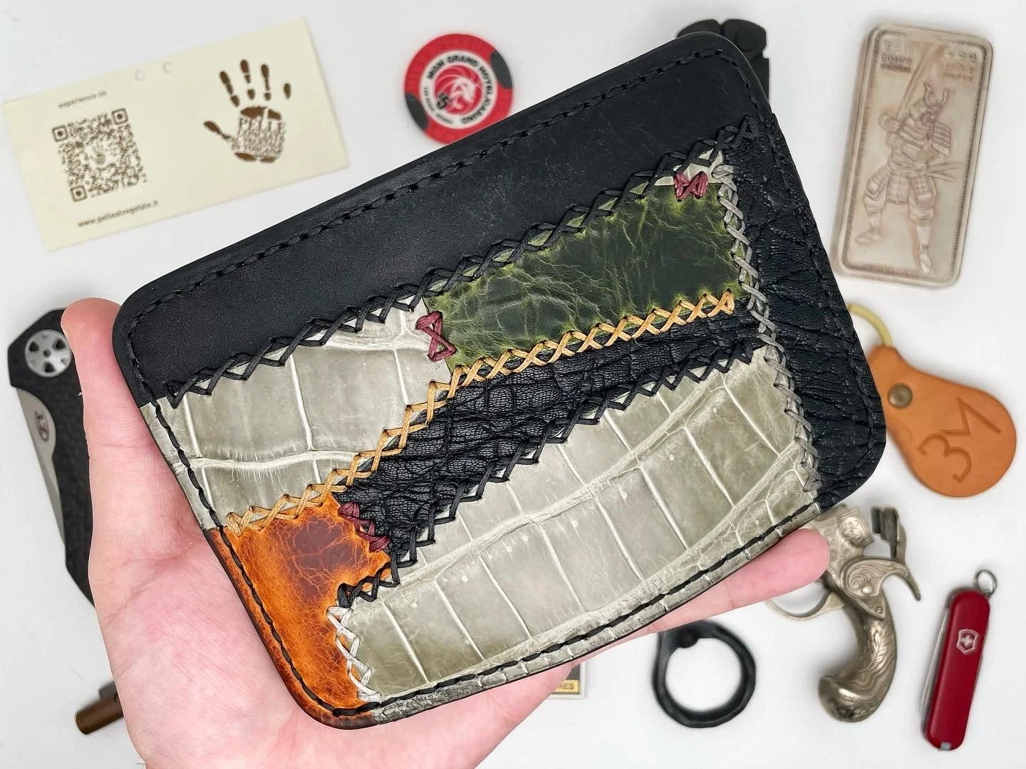A patchwork wallet made of various textured leathers and fabrics, being held in hand, with other miscellaneous items including a Swiss Army knife, keychain, metal keyring, and various small objects on a white surface in the background.