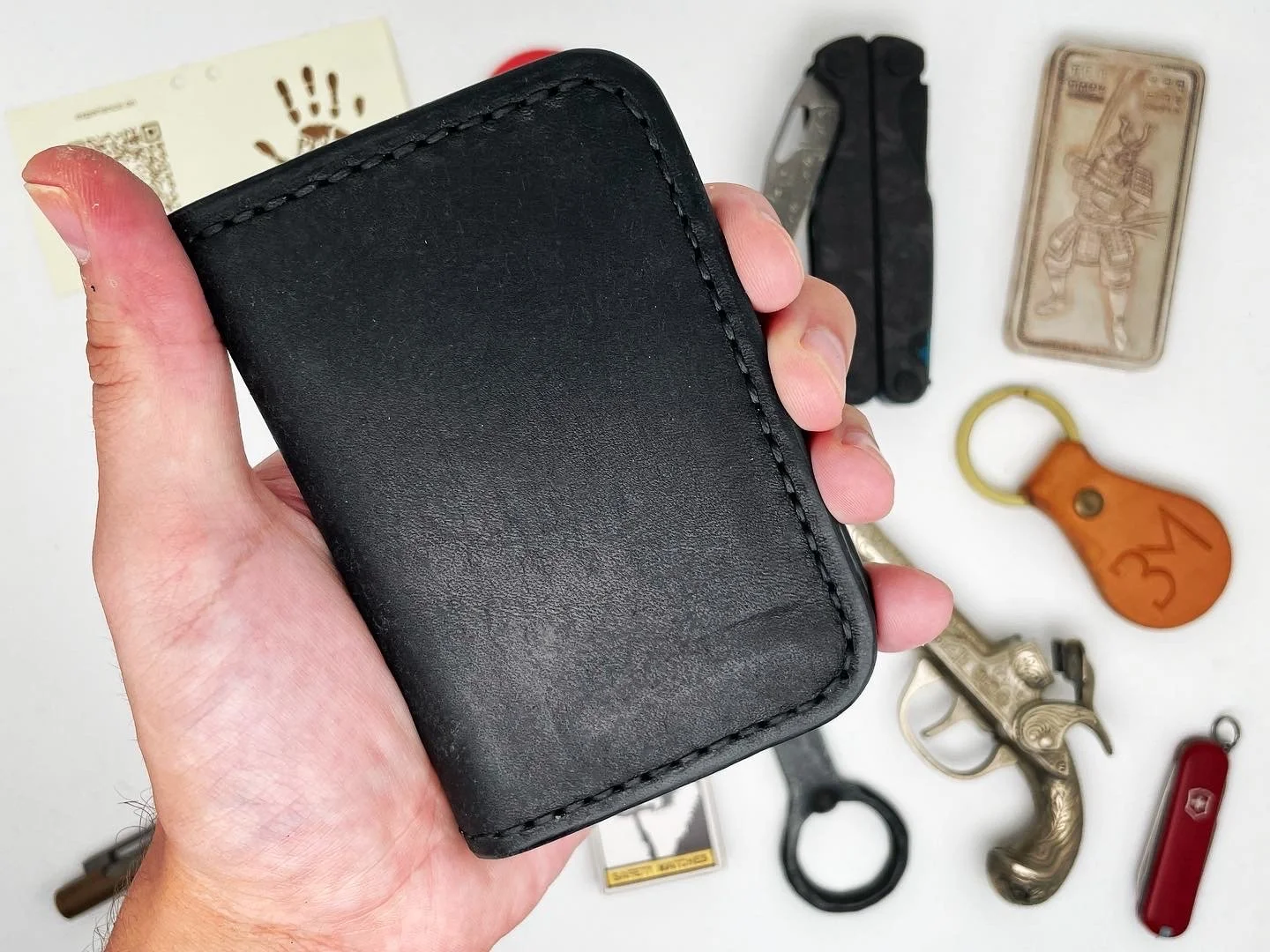 A person's hand holding a black leather wallet with various small objects laid out on a white surface behind it, including a finger-shaped keychain, a small knife, a toy gun, a Swiss Army knife, a wooden keychain, a small pair of handcuffs, a framed 