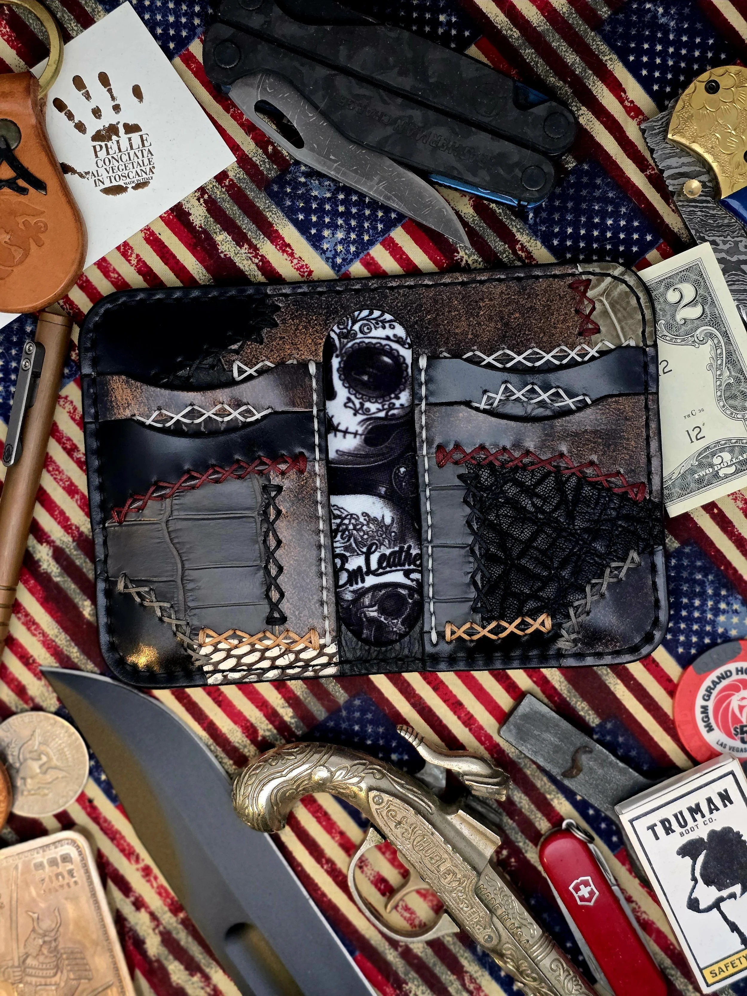 A collection of knives, a dollar bill, a Swiss army knife, a gold handcuff, a gold revolver, coins, and a trader sticker on a red, white, and blue American flag-patterned fabric.