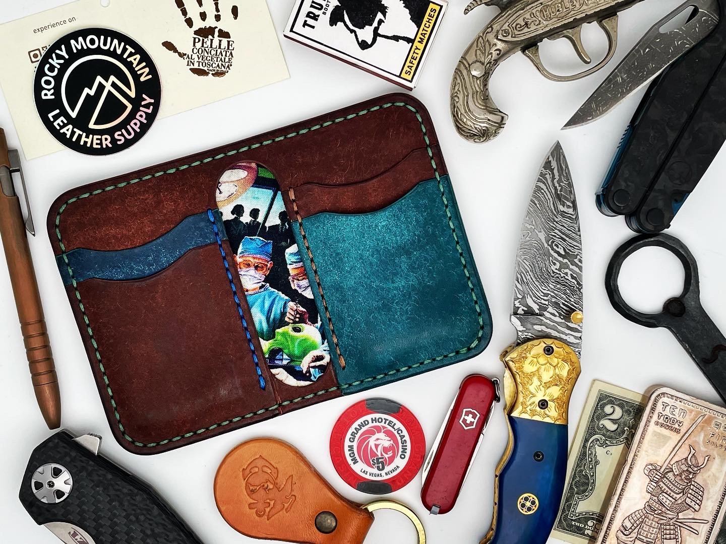 A collection of everyday items on a white surface including a brown and blue leather wallet with a photo of children wearing masks, several knives with various designs, a multi-tool, a small black flashlight, currency bills, a poker chip, a Swiss arm