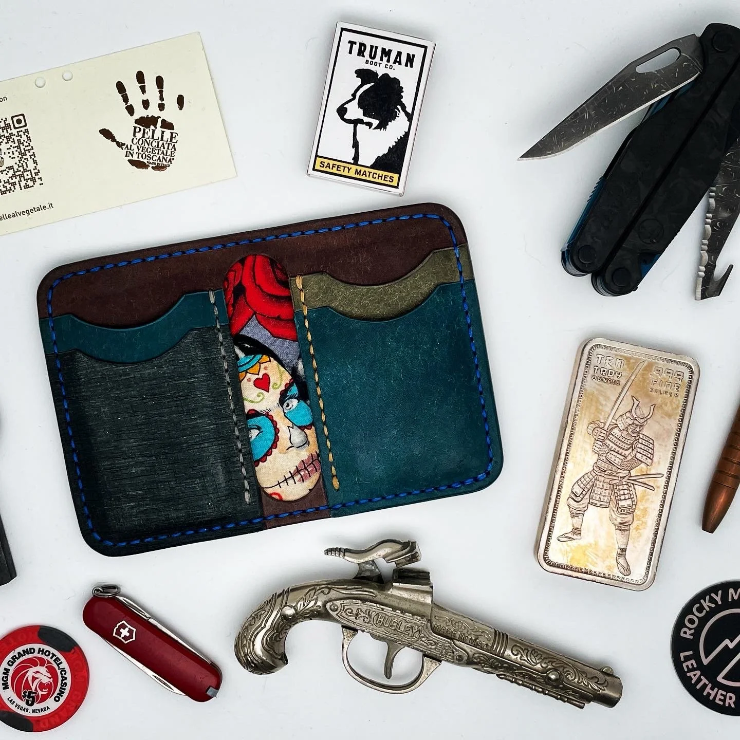 Collection of miscellaneous items including a colorful leather wallet, a folding knife, a decorative metal pistol, a red Swiss Army knife, a small metal pin with lion logo, a matchbox with lion image, a pack of safety matches, and various other small