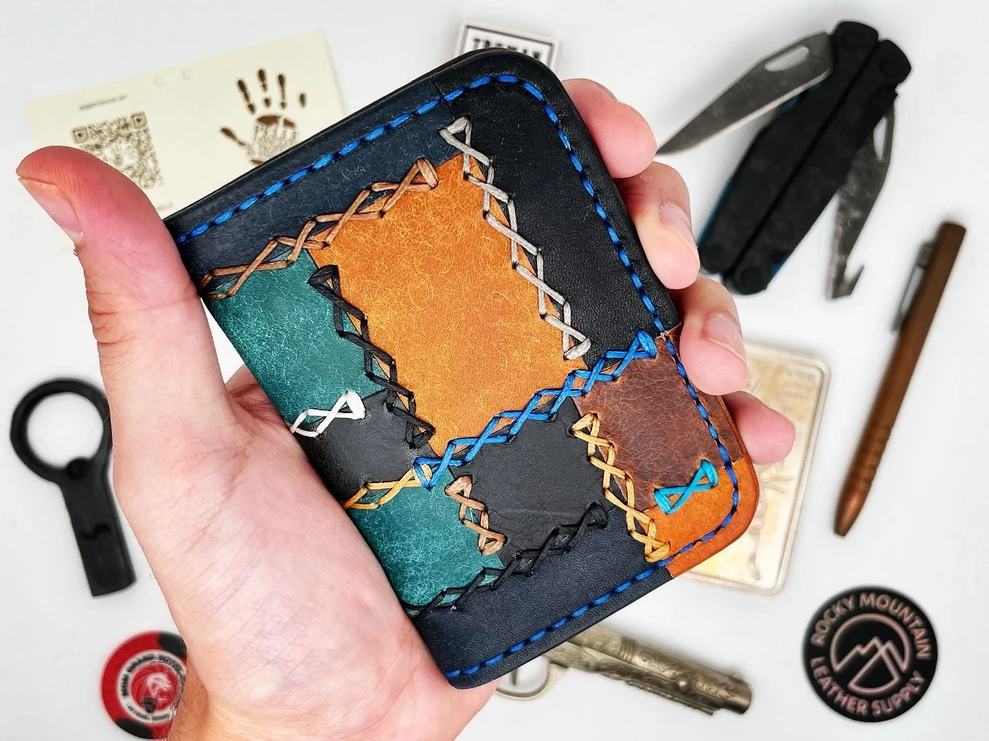 Hand holding a colorful leather wallet with patchwork design and stitched border, surrounded by various tools and stickers on a white surface.