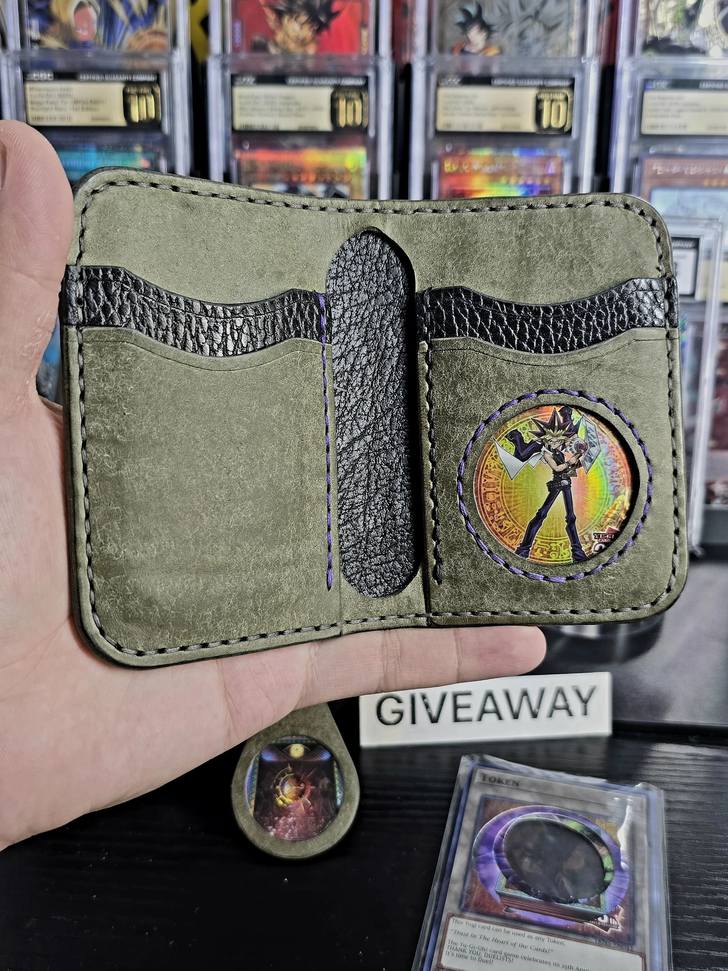A hand holding an open Yu-Gi-Oh! themed wallet with cards inside, set against a background of card storage boxes with a 'GIVEAWAY' sign and a holographic card holder nearby.