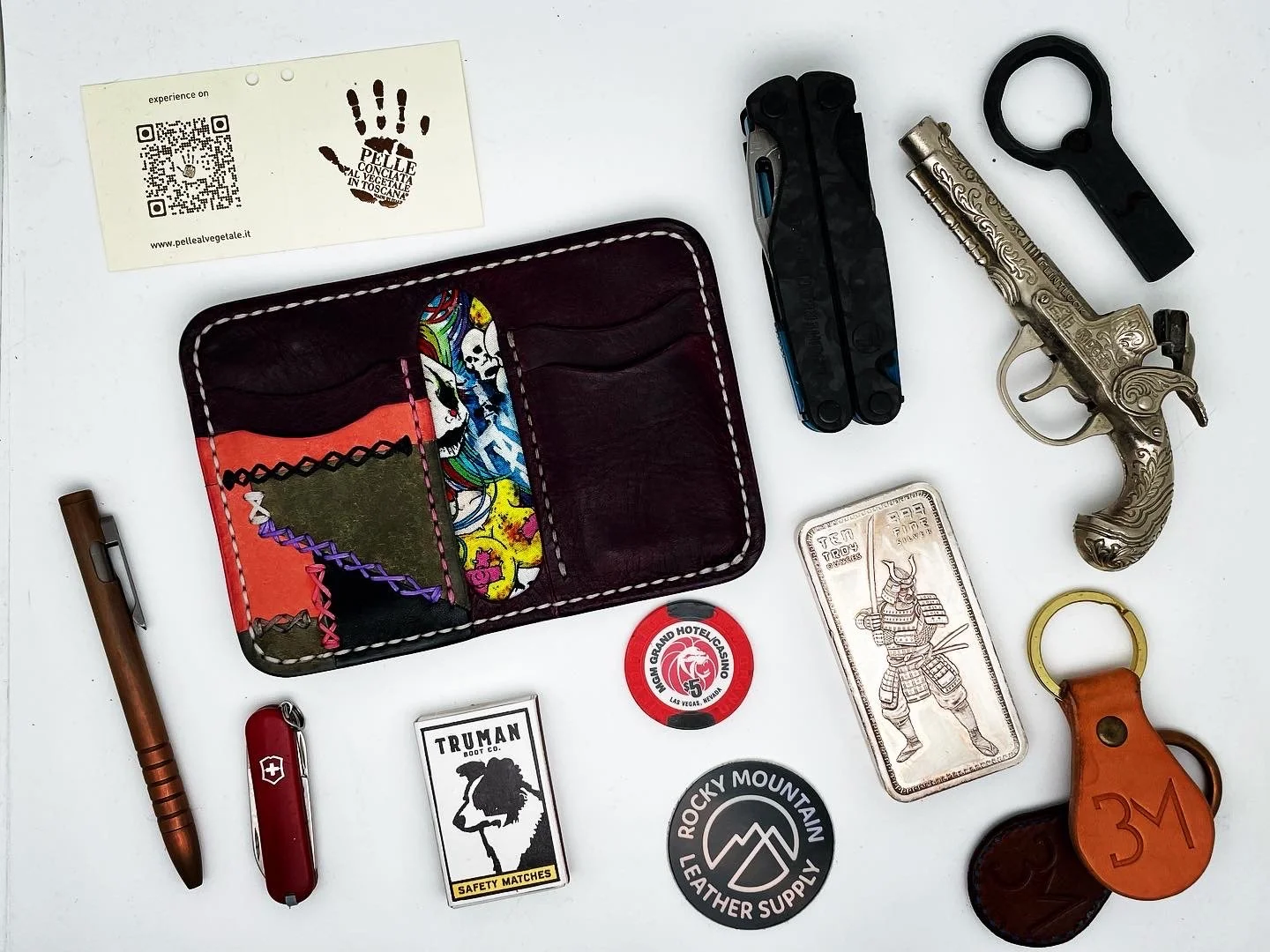 Collection of various items including a colorful leather wallet, a tactical black multitool, a vintage silver revolver, a black key ring, a copper-colored flashlight, a red Swiss Army knife, a pack of safety matches, a round poker chip, a silver bar 