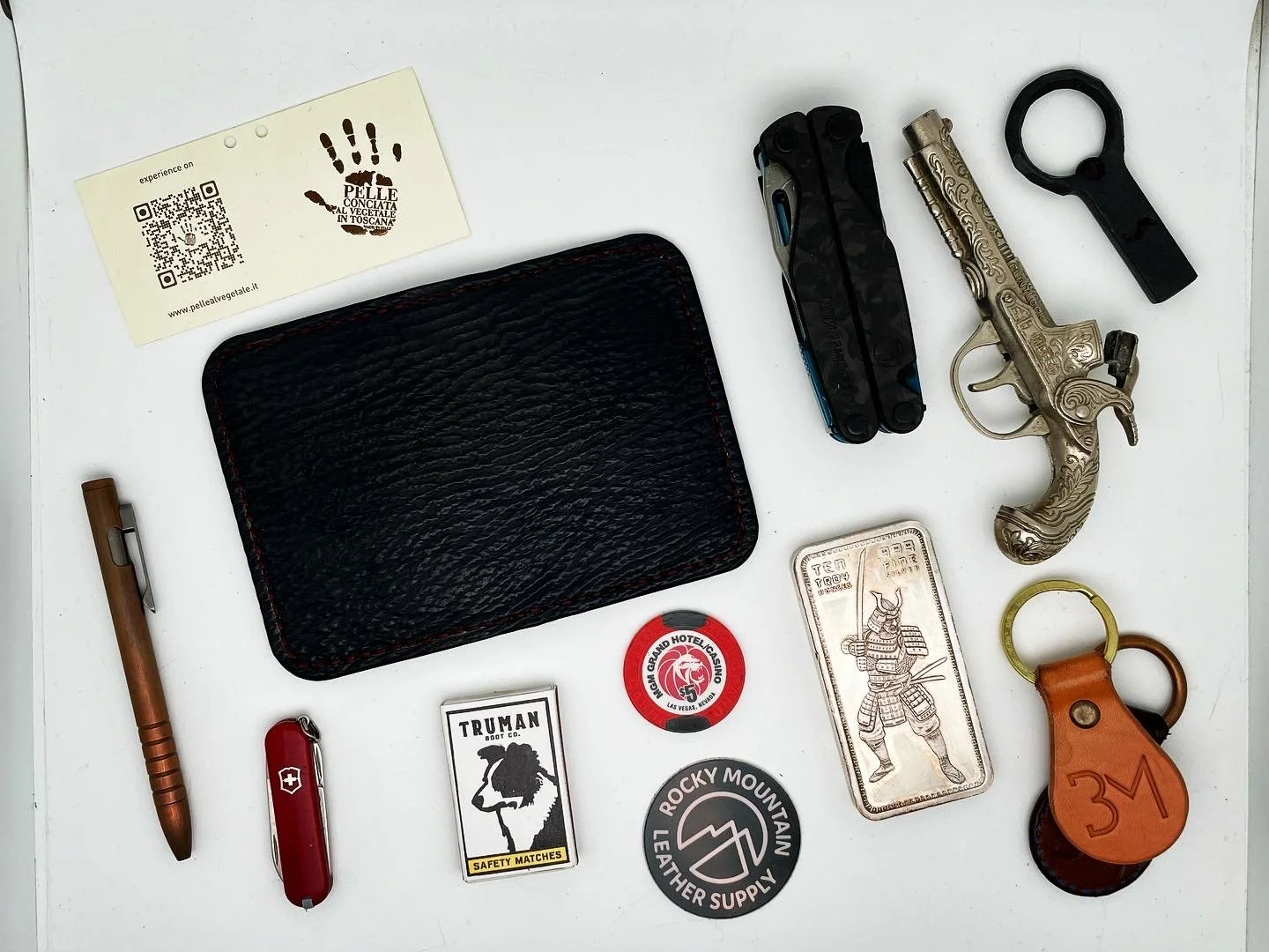 Collection of everyday carry items on a white surface, including a black leather wallet, a gold engraved revolver-shaped lighter, a wooden pen, a red Swiss Army knife, a small rectangular dog tag, a black keychain, a silver bar with a samurai figure,