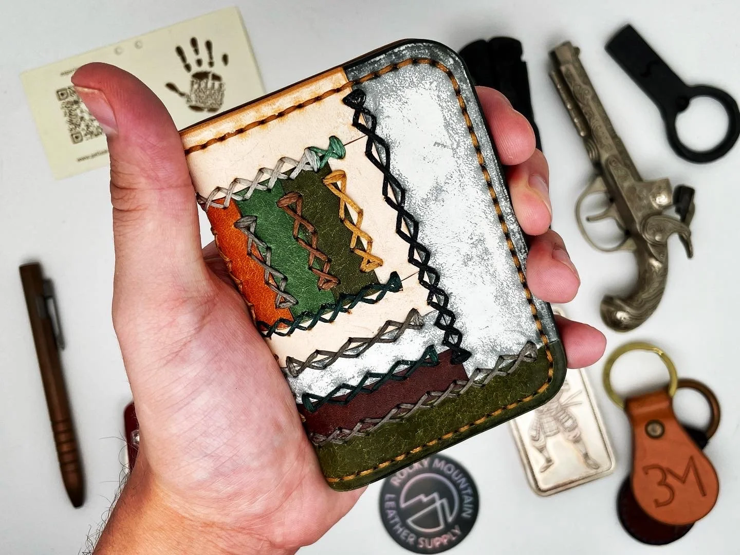 Close-up of a person's hand holding a small leather wallet with orange, green, and brown sections, stitched with colorful thread. On a white surface in the background are various items, including a silver revolver, a black knife, a keychain with the 