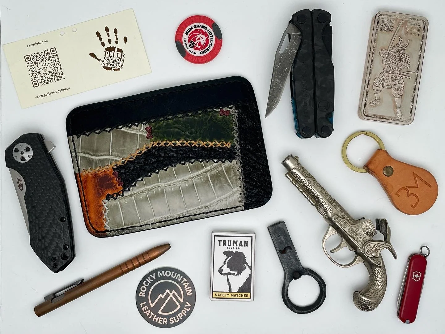 Collection of everyday carry items on a white surface, including folding knives, a pistol, a leather and glass wallet, a keychain, a mini lighter, a pen, patches, a safety match book, and a business card.