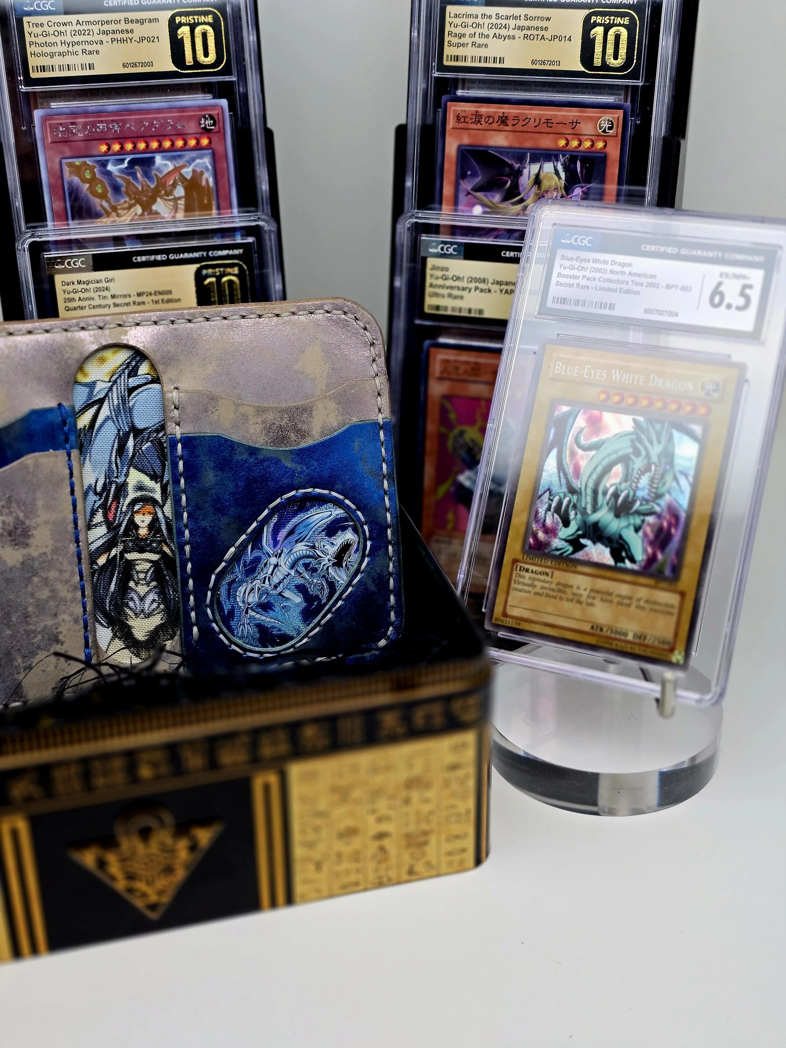 Yu-Gi-Oh! trading cards in display cases and a card wallet with a Dragon and a Wolf design. The display cases show graded and authenticated Yu-Gi-Oh! cards, including a Blue-Eyes White Dragon card graded 6.5.