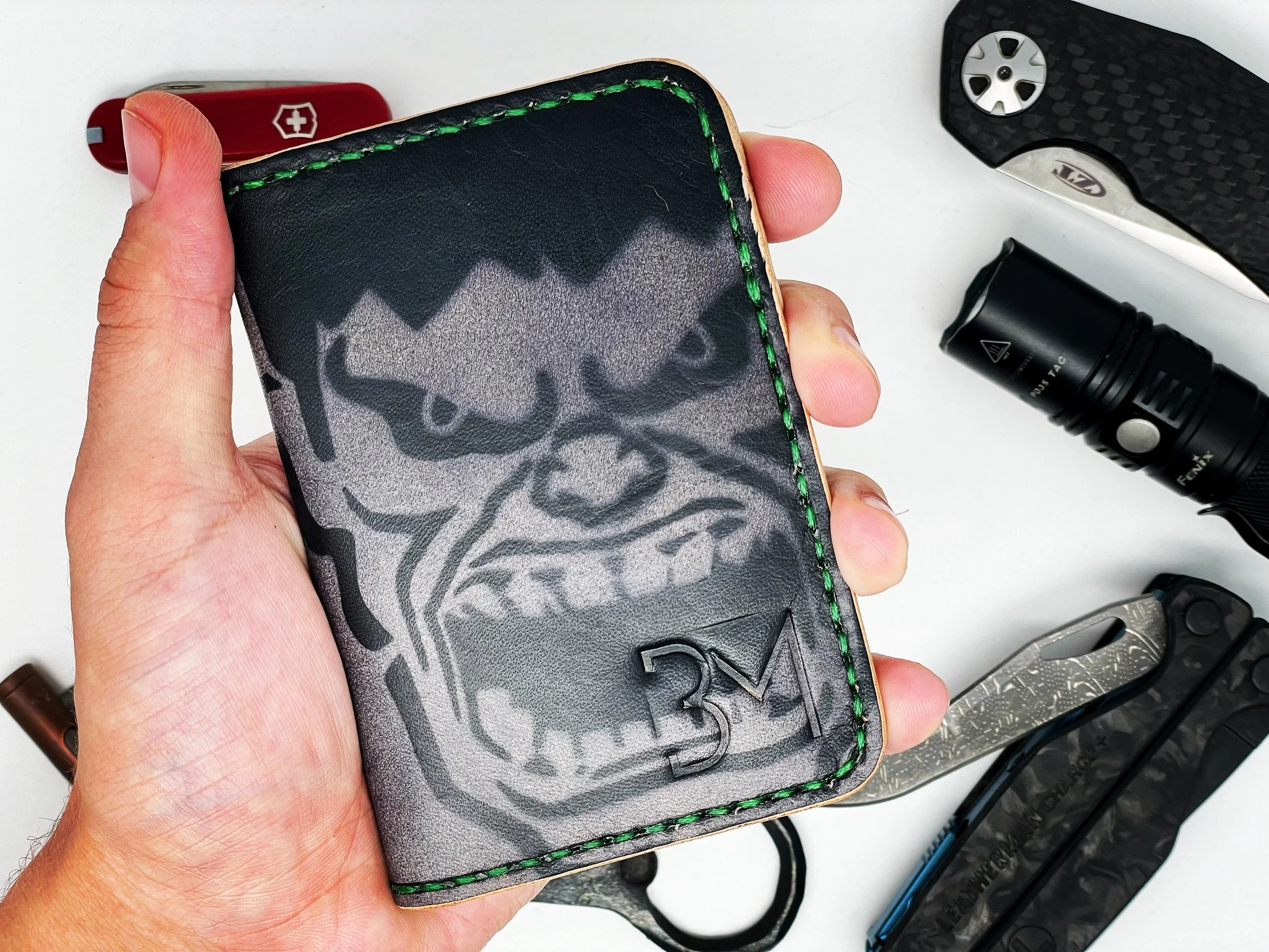 A hand holding a black leather wallet with a gray dragon graphic and green stitching, surrounded by pocket knives, a flashlight, and other tools on a white surface.