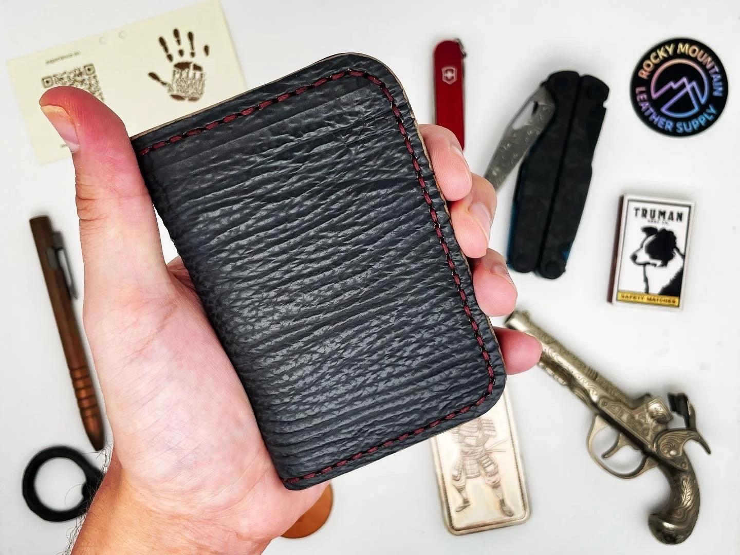 Hand holding a black leather wallet with red stitching, surrounded by various objects including a Swiss Army knife, a vintage pistol, a badge, a matchbox, a cigar, and other small items on a white surface.