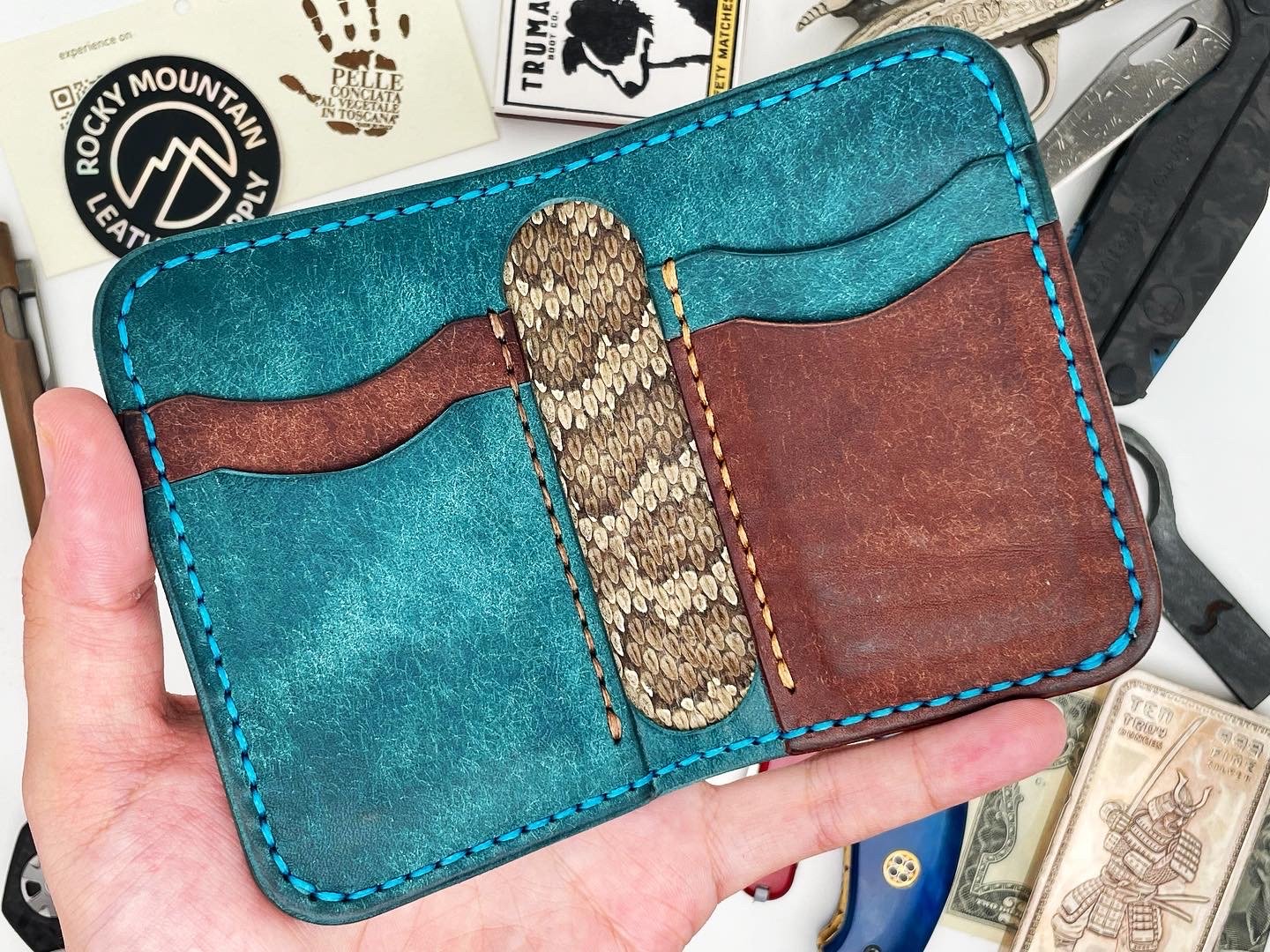 Hand holding a colorful leather wallet with card slots, featuring teal, brown, and snake print designs, on a cluttered surface with stickers, knives, and paper currency.